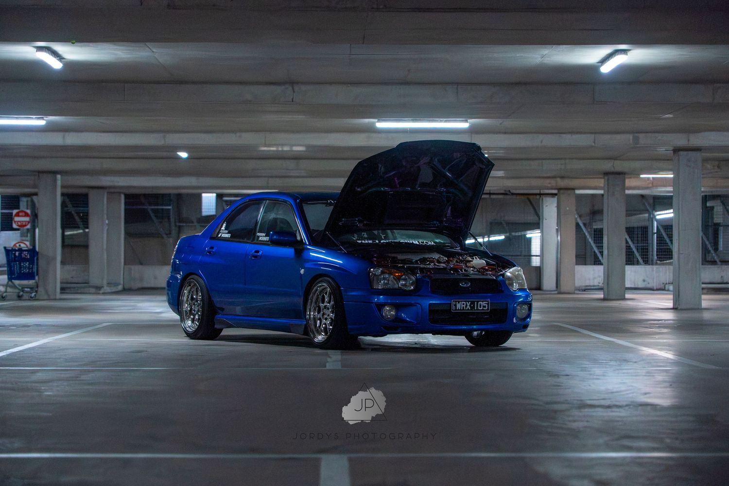 Modified blue Subaru Impreza with popped hood displayed in underground parking structure at night.