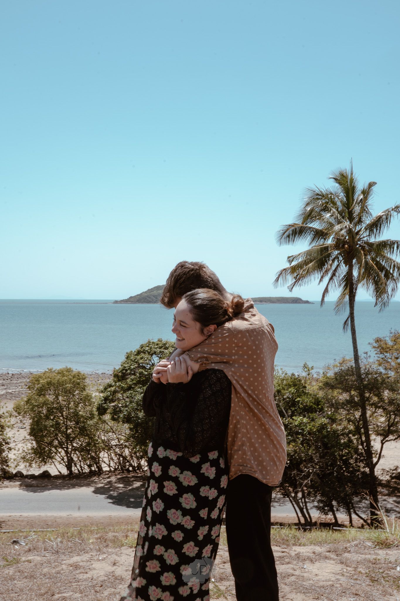 Two figures embrace while enjoying an oceanside view with palm trees and blue skies.