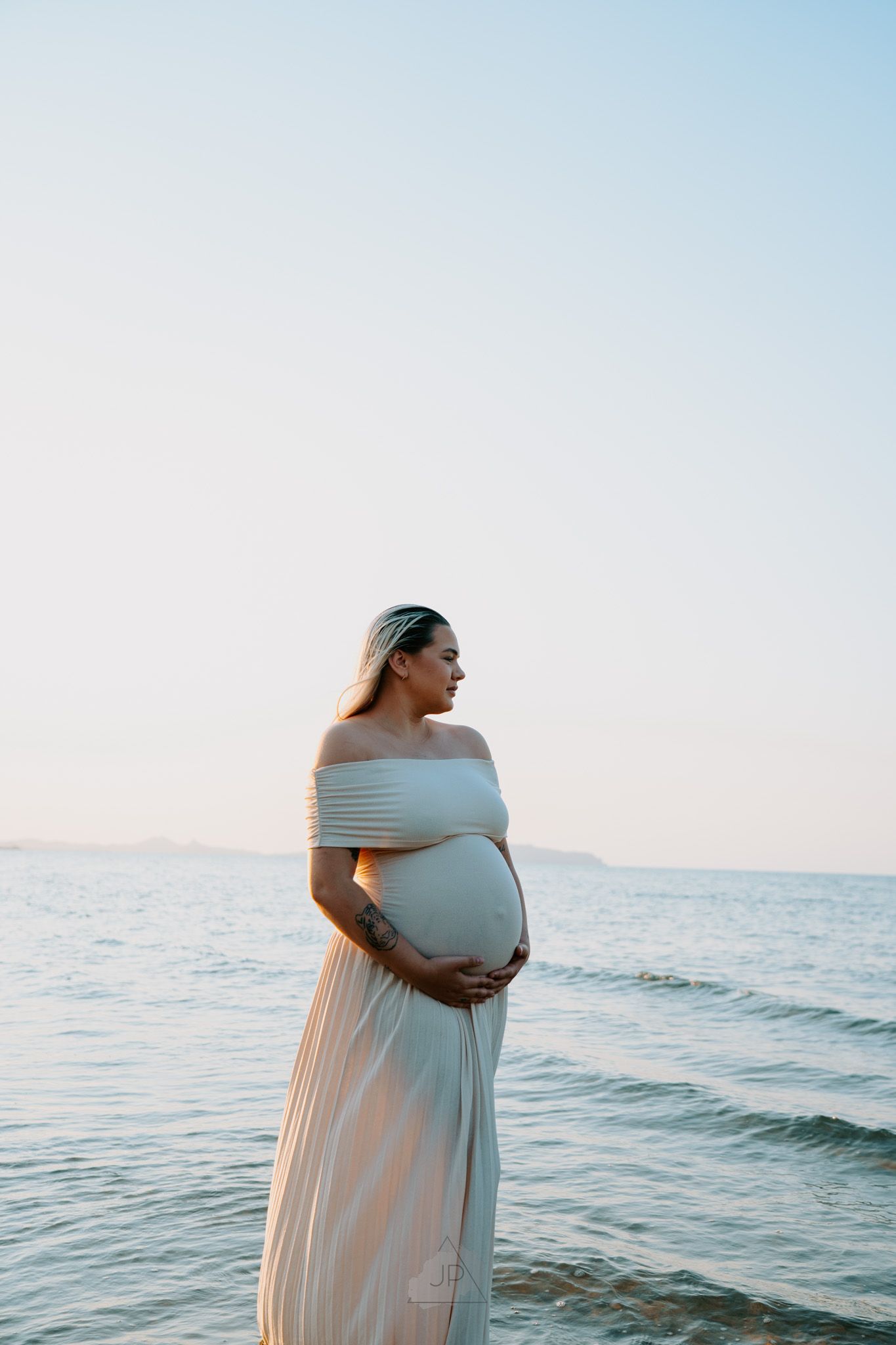 Pregnant woman in flowing white dress stands by ocean at sunset holding her baby bump in four progressive poses.