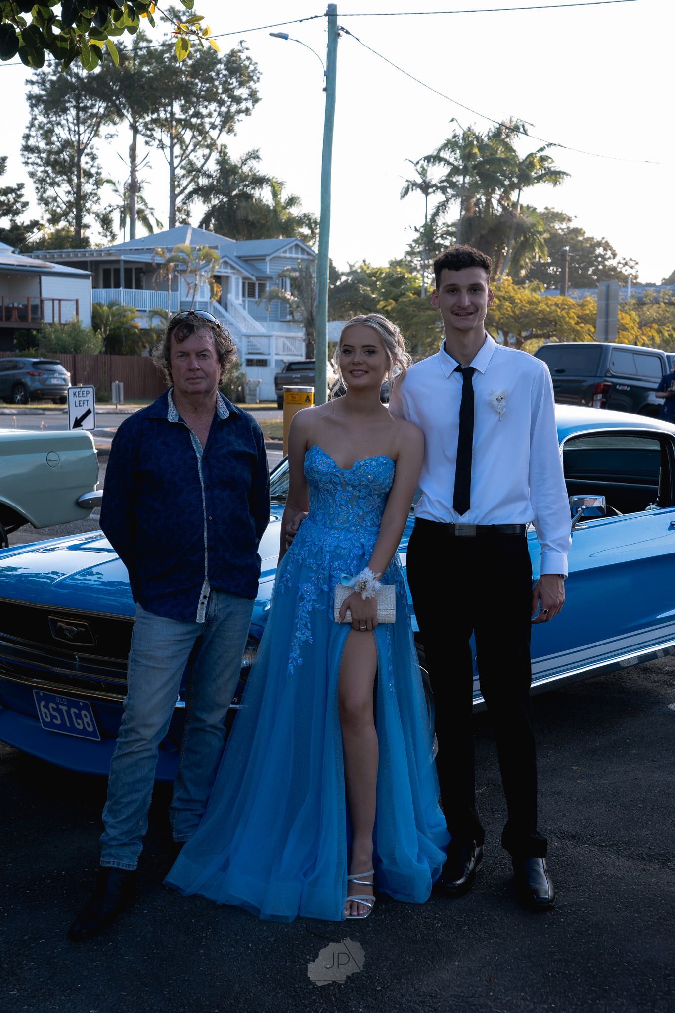 A group in formal attire poses beside a classic blue car at dusk during a special occasion.