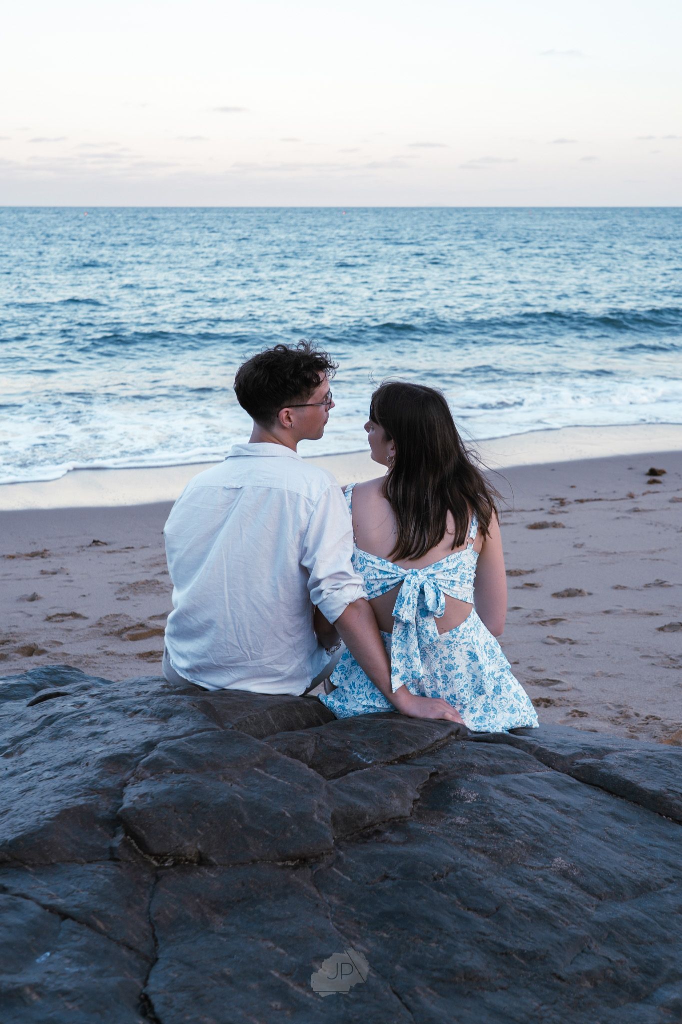 A couple sits together on a sandy beach at sunset watching the ocean waves roll in.