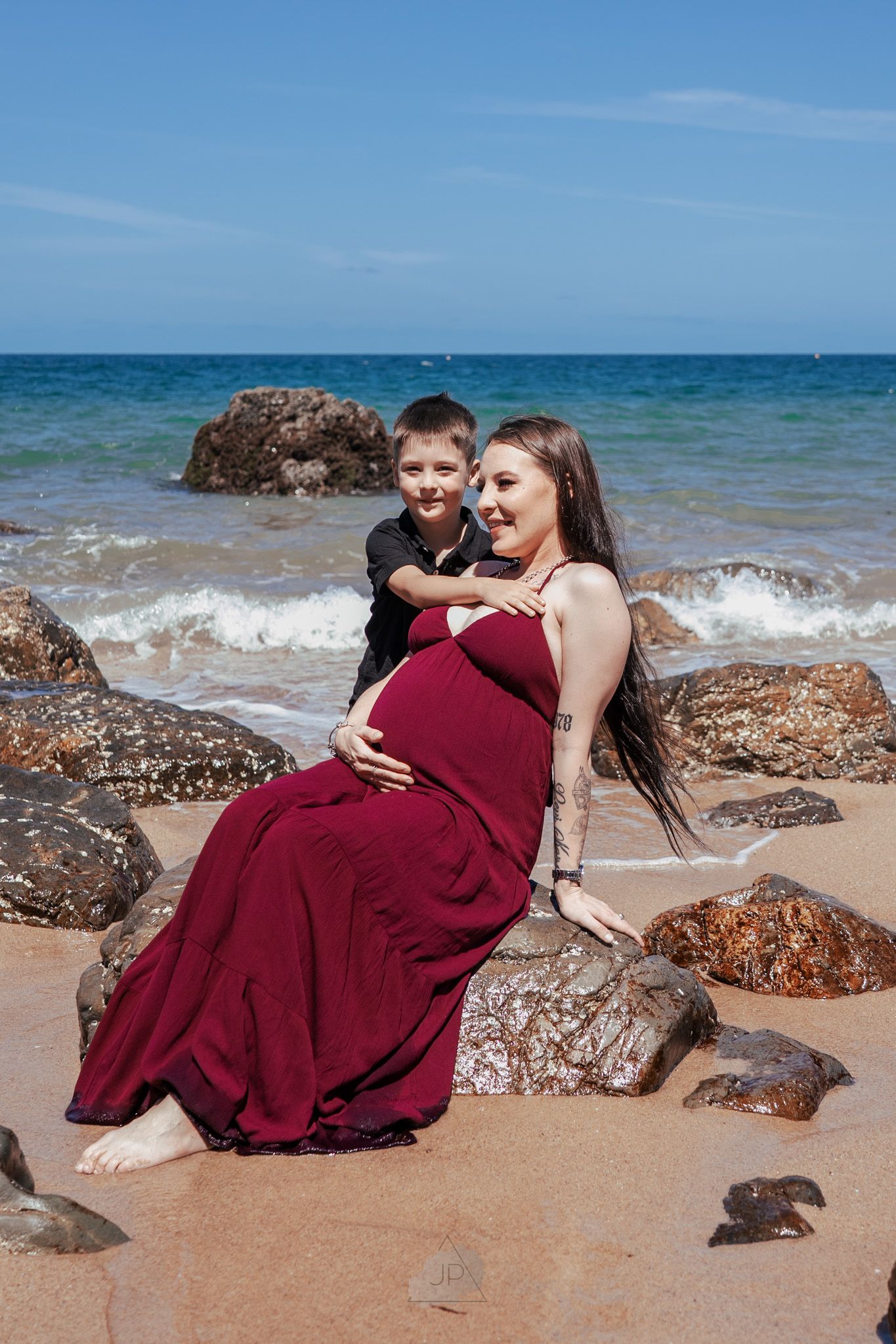 A romantic couple in burgundy attire embraces on a sandy beach with ocean waves and rocks in the background.