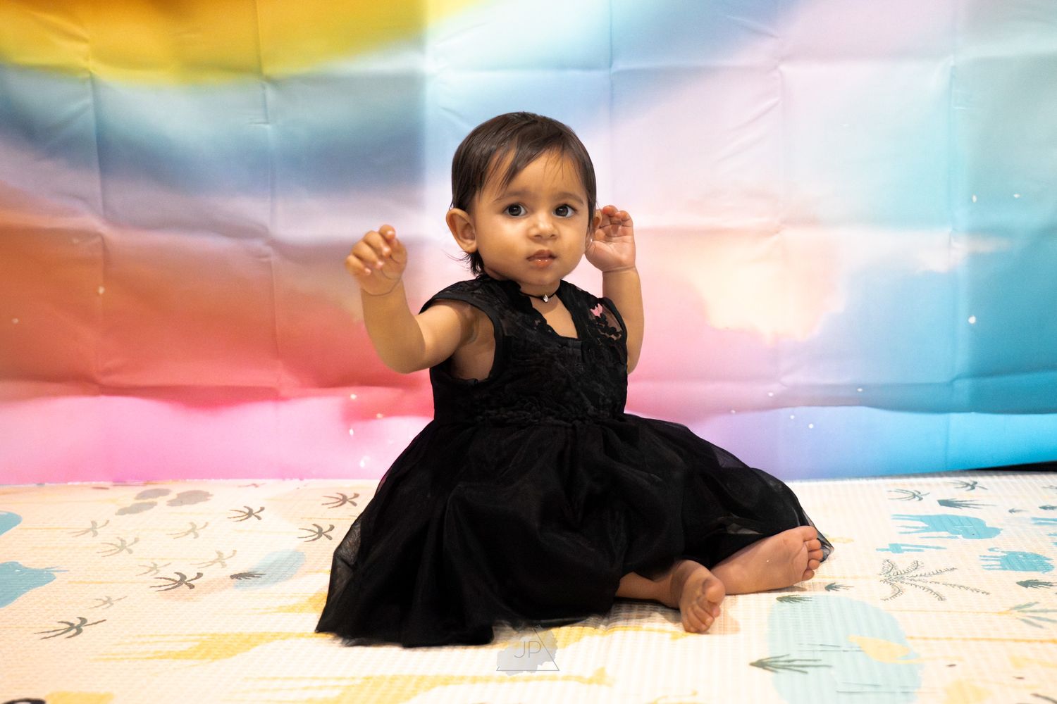 A baby in a black tutu dress crawls on a colorful pastel backdrop during a photo session.