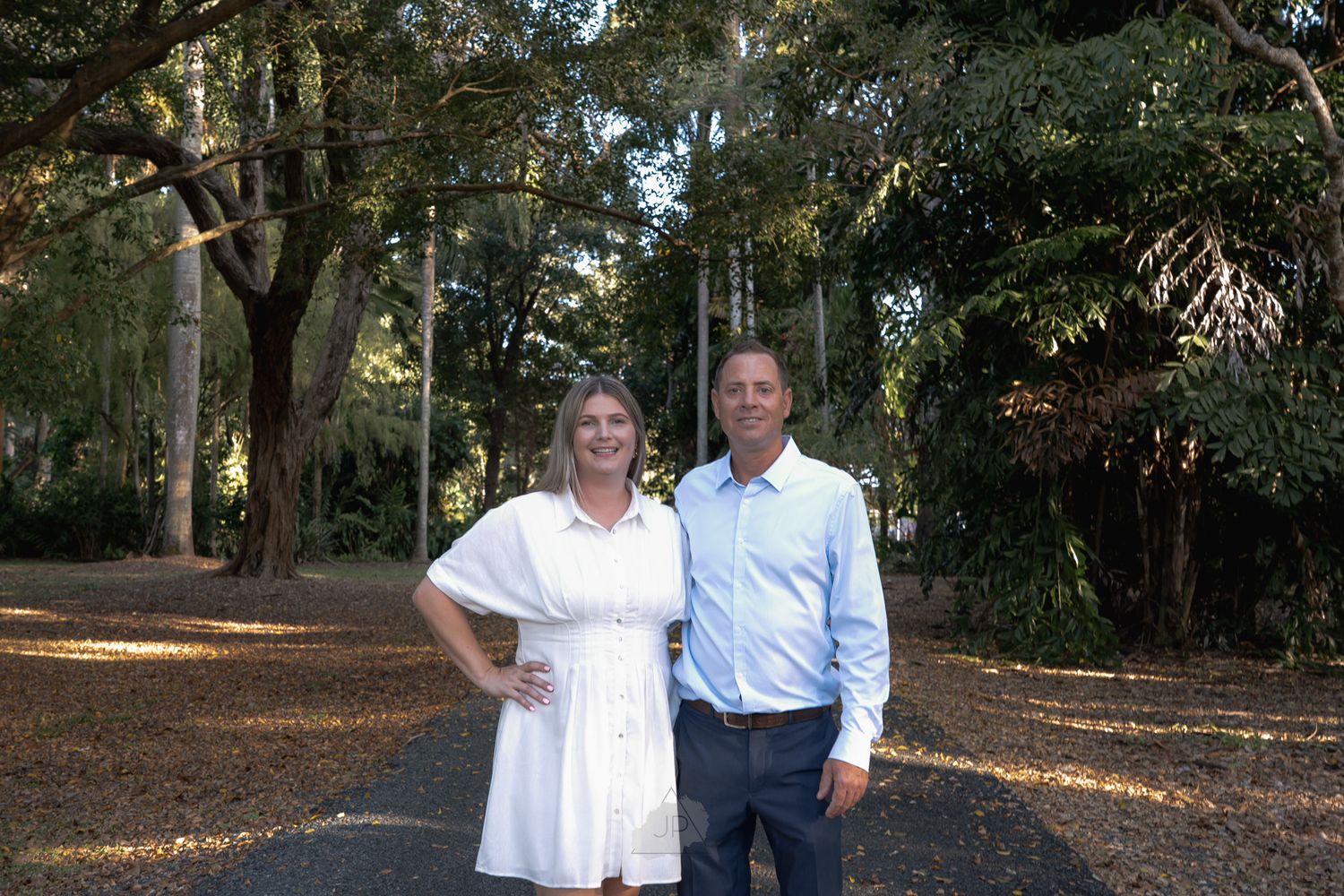 Two people stand together on a path surrounded by trees in a wooded outdoor setting.