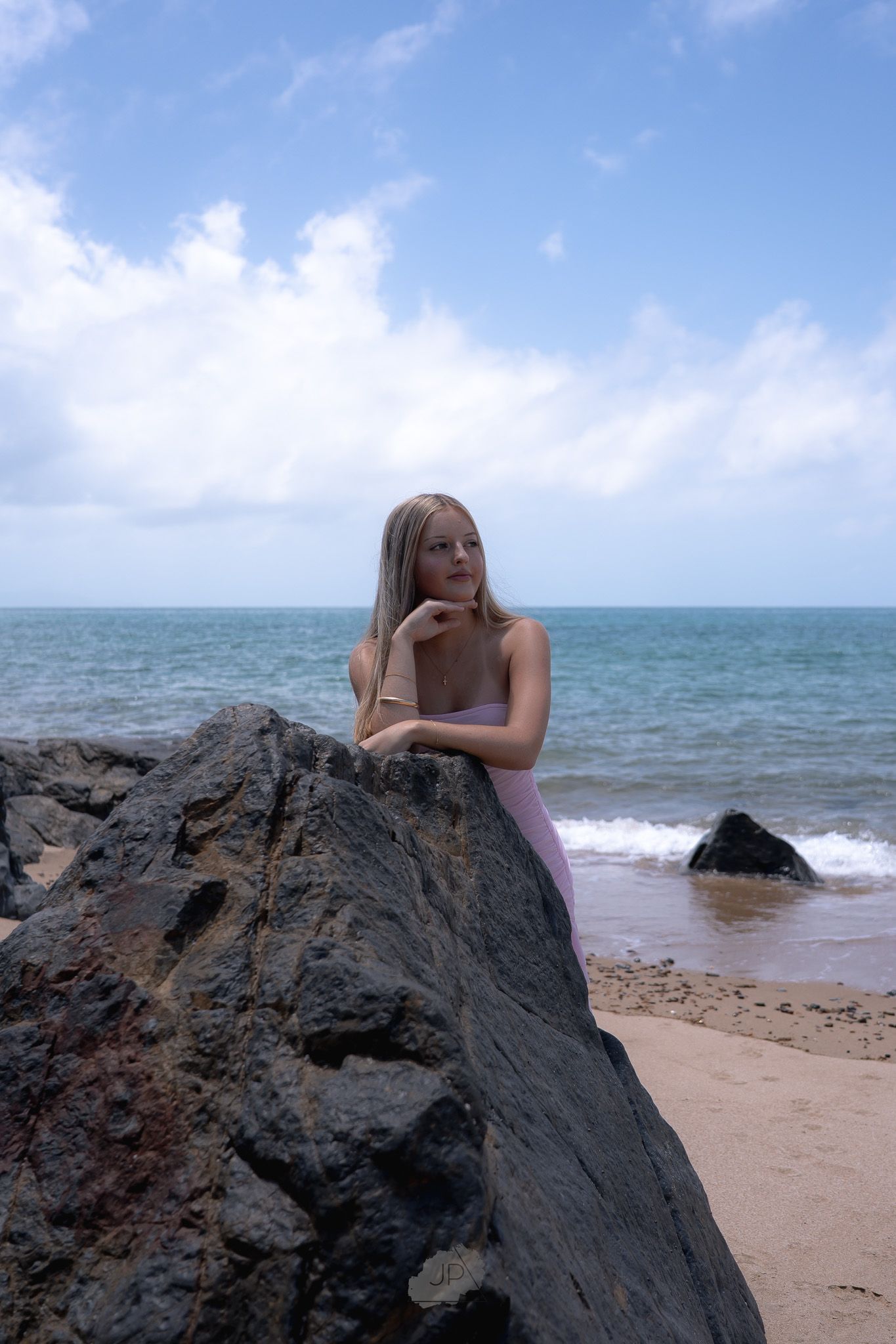 A person lounges on dark volcanic rocks at a tropical beach with turquoise waters and white sand under blue skies.