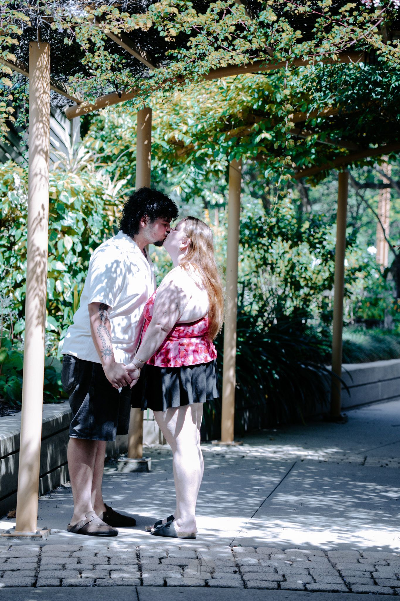 A couple shares a romantic moment under a tree-lined path in a park setting.