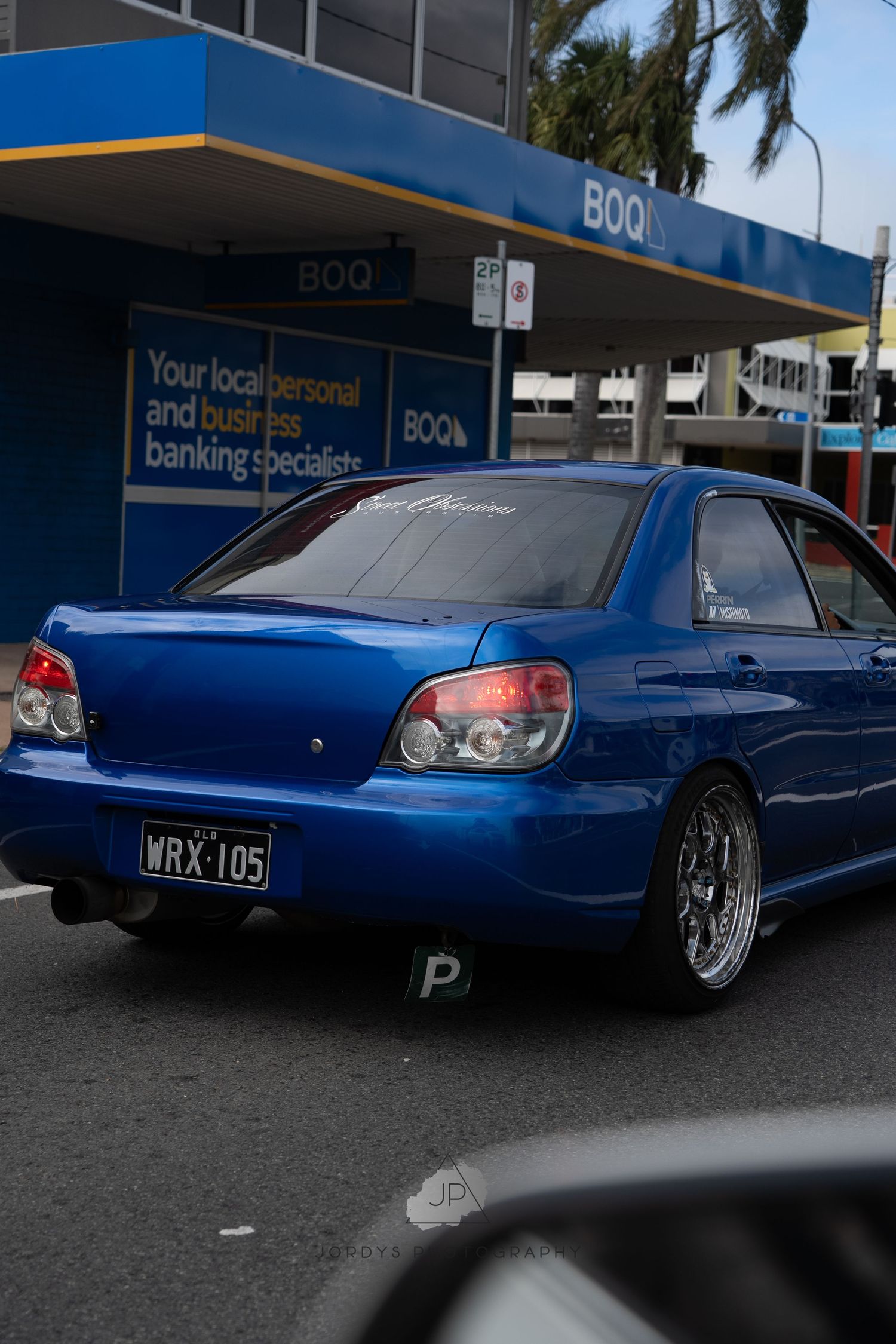 A sleek blue modified car with custom wheels parked in front of a bank building.