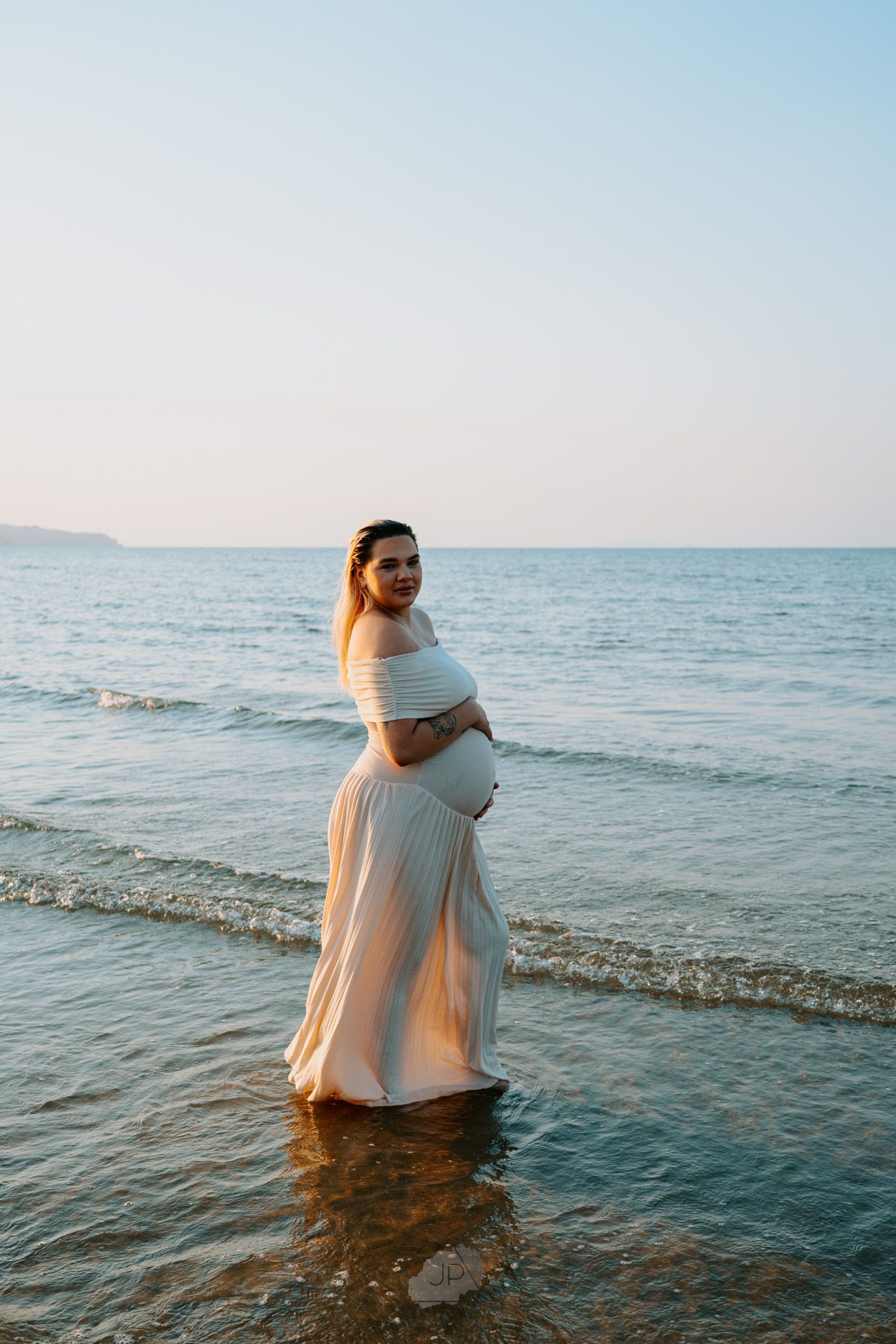 A pregnant figure in a flowing white dress stands in shallow ocean water at sunset with gentle waves rolling in.
