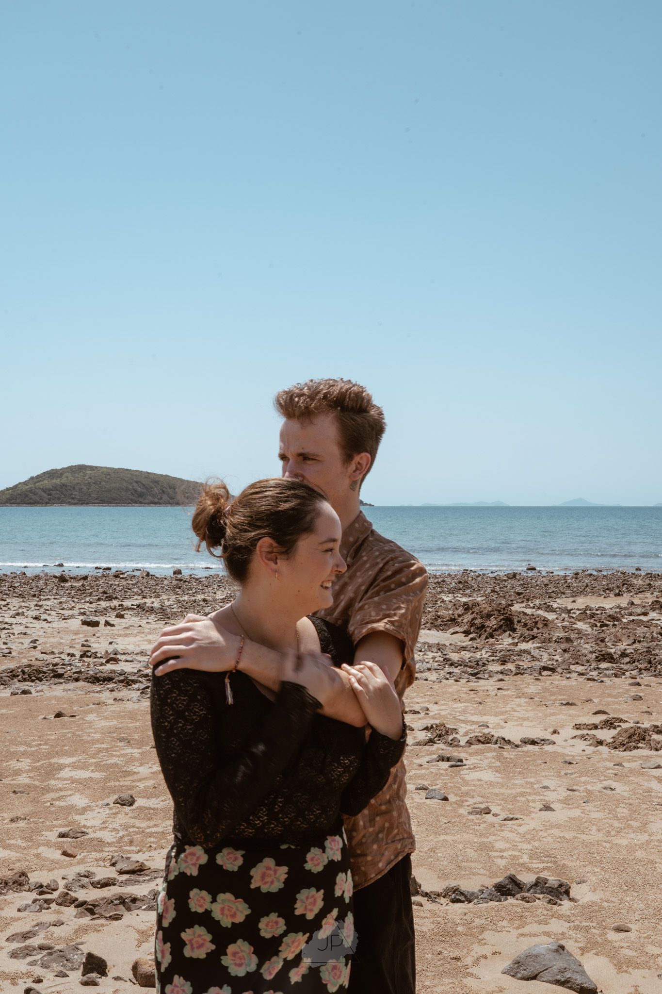 A couple embraces and kisses on a rocky beach shoreline with ocean waves visible in the background.