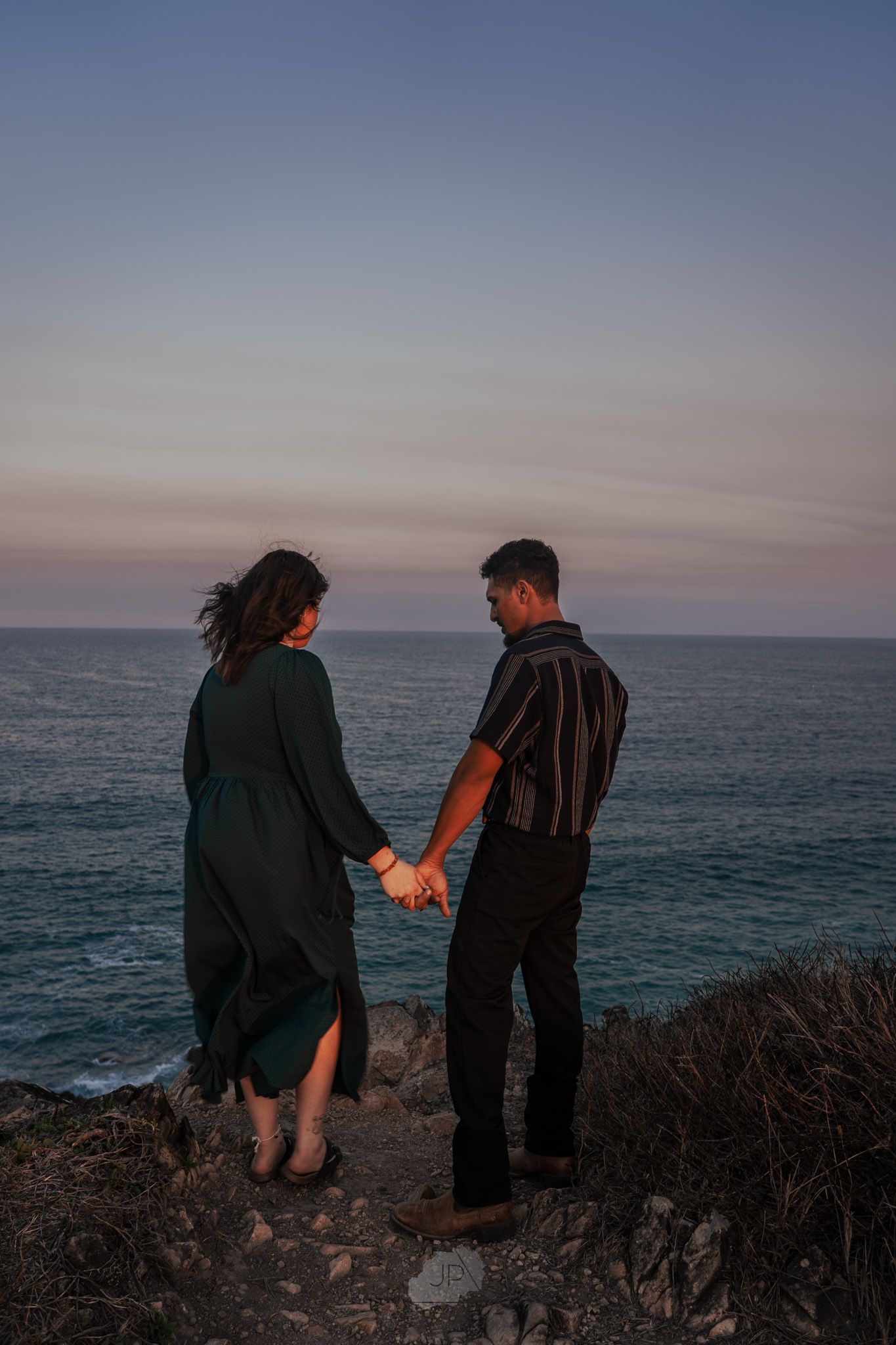 Two people stand together on a rocky coastal cliff overlooking the ocean at sunset.