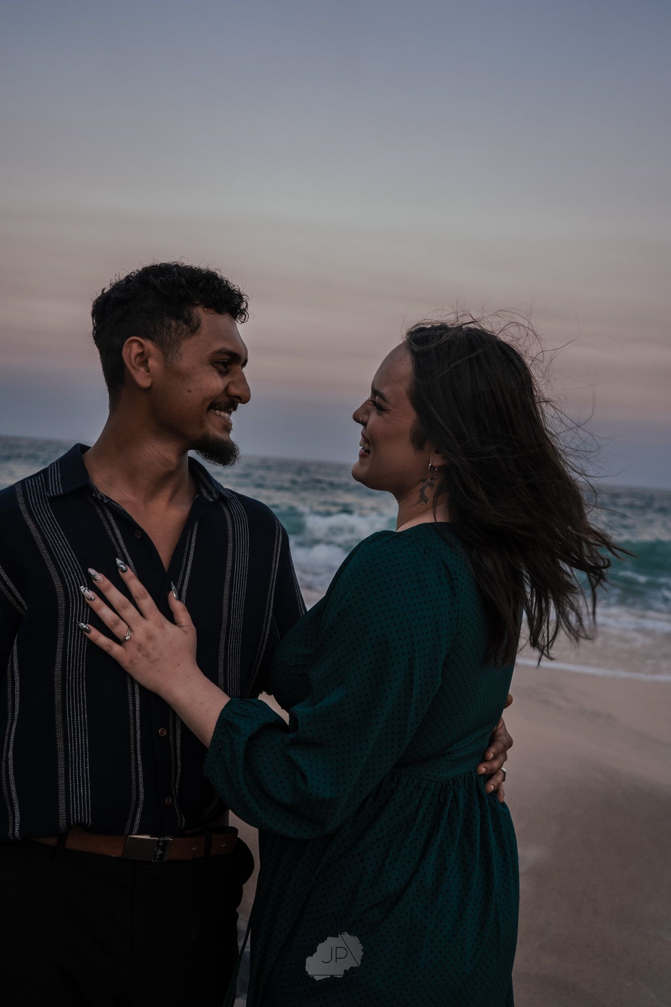 A romantic couple shares laughter and embraces while walking along a misty beach at sunset.