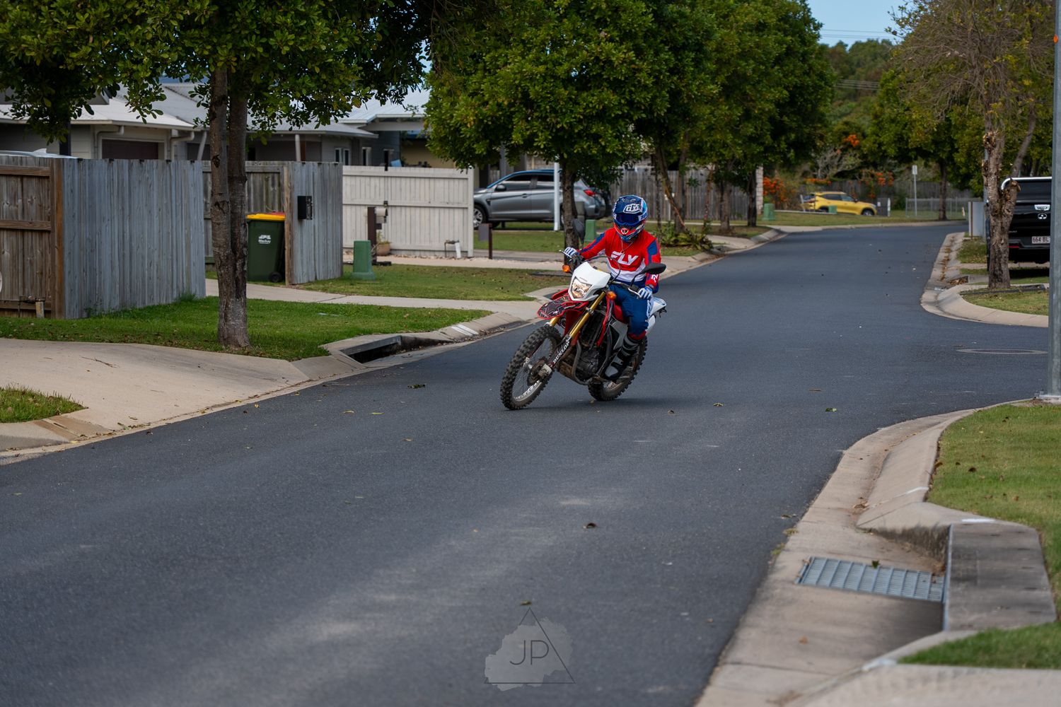 Wide-angle view shows motorcycles and cars parked on residential suburban streets with green trees and lawns.