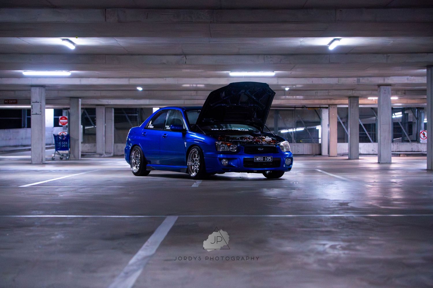 A blue sports car parked in an underground concrete parking garage with fluorescent lighting overhead.