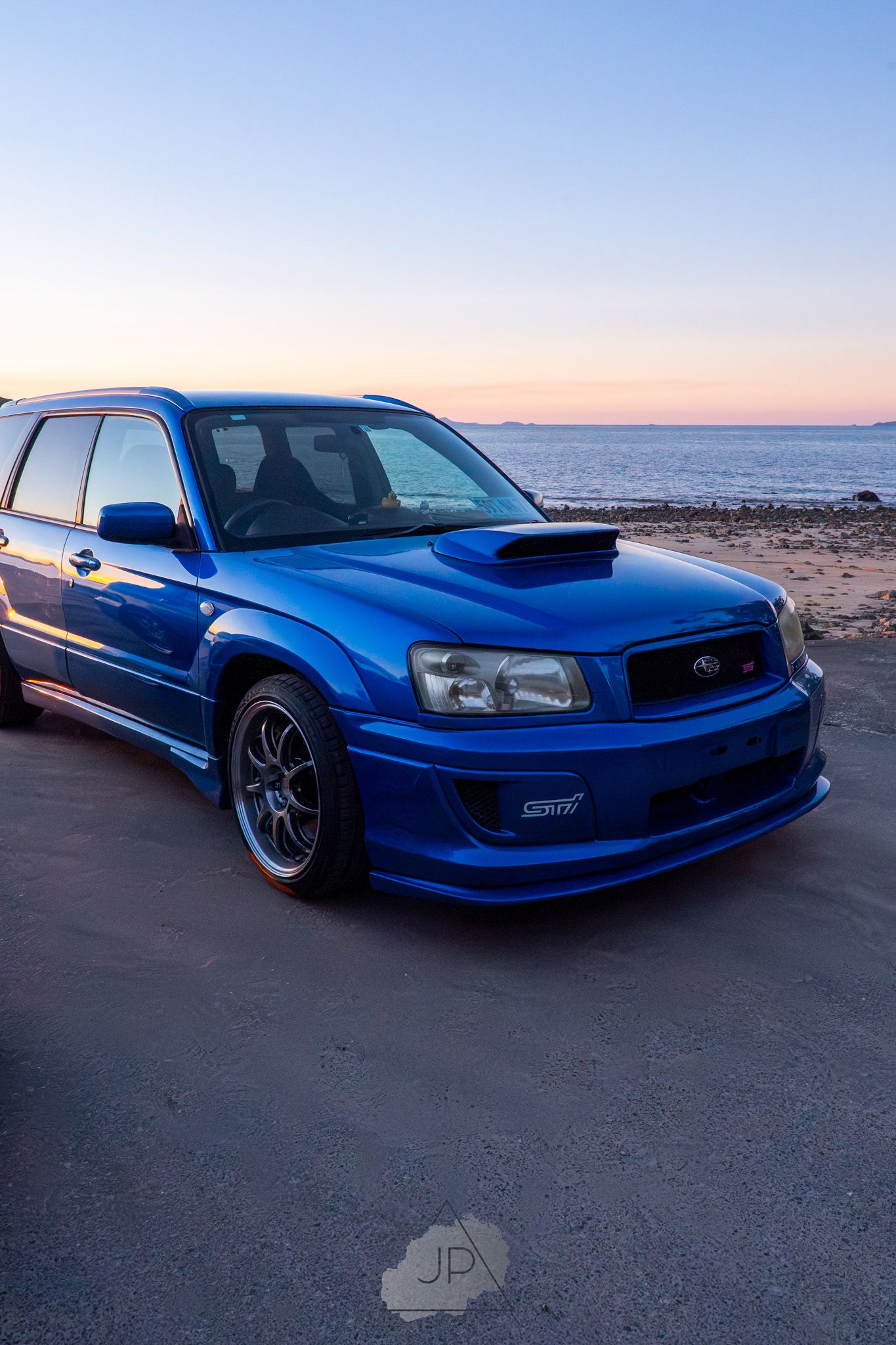 Customized blue Subaru Forester STI sits parked at sunset along a scenic coastal shoreline.