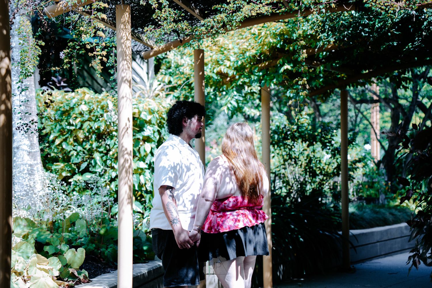 A group of people walk through a lush green garden pathway surrounded by tall trees and foliage.