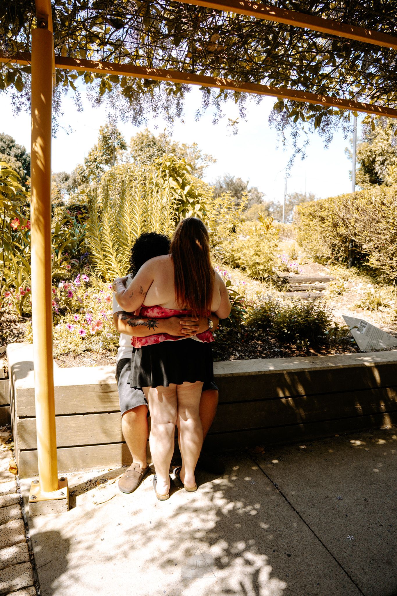 Two people sitting on a wooden bench under a shaded outdoor structure on a sunny day.