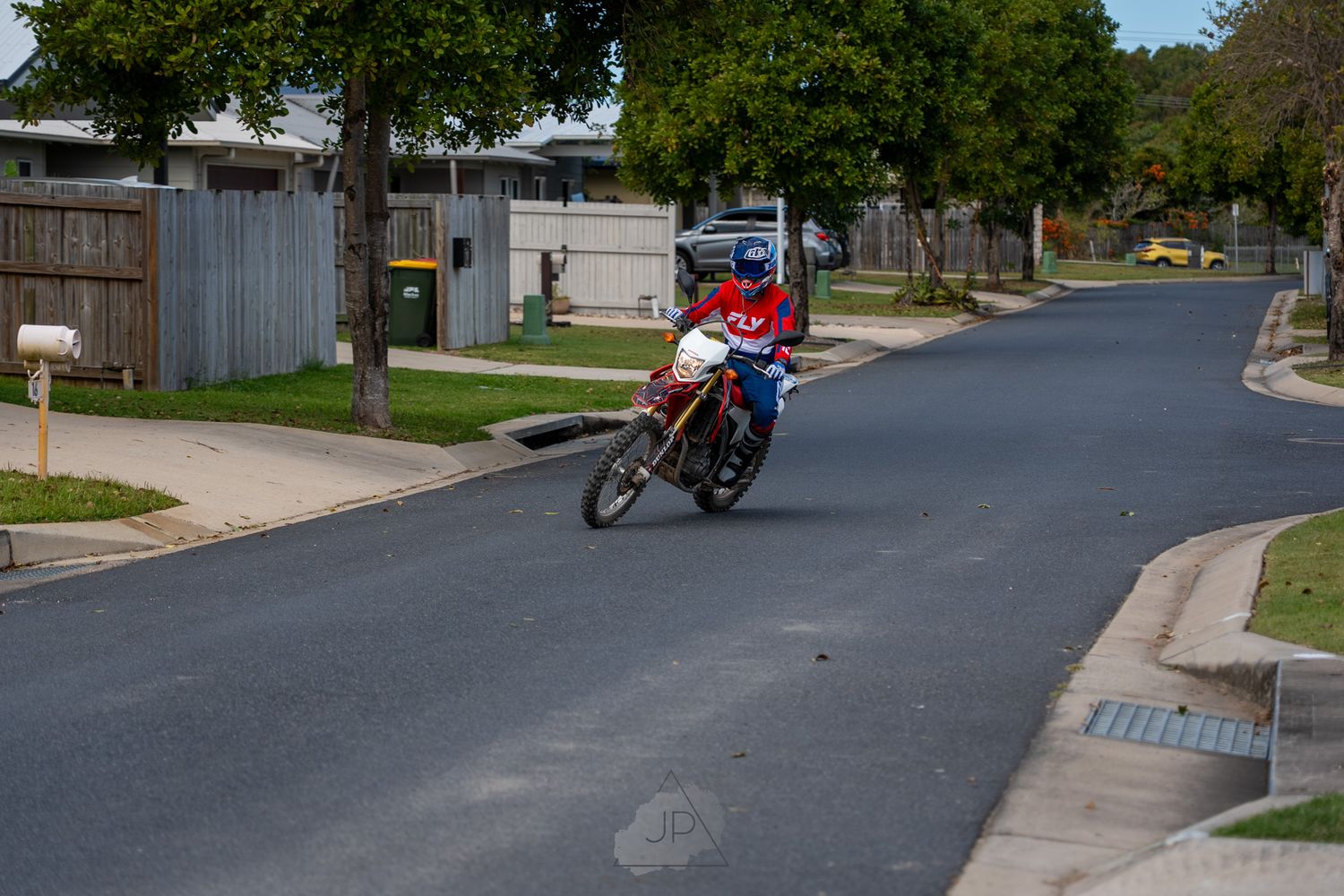 A motorcyclist rides a red dirt bike on a suburban street lined with trees and fences.