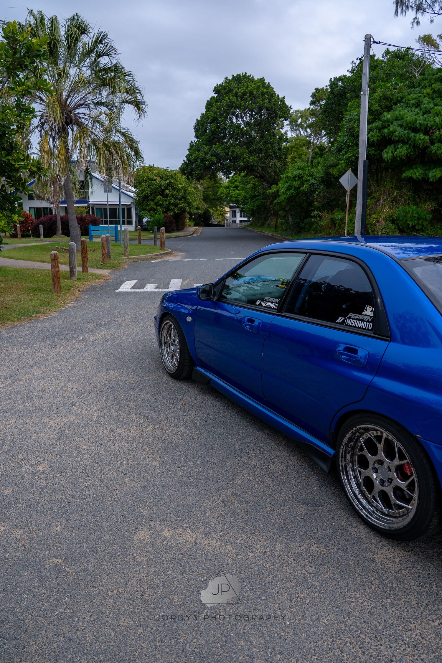 A blue Subaru Impreza WRX with custom wheels parked on a residential street lined with palm trees and greenery.