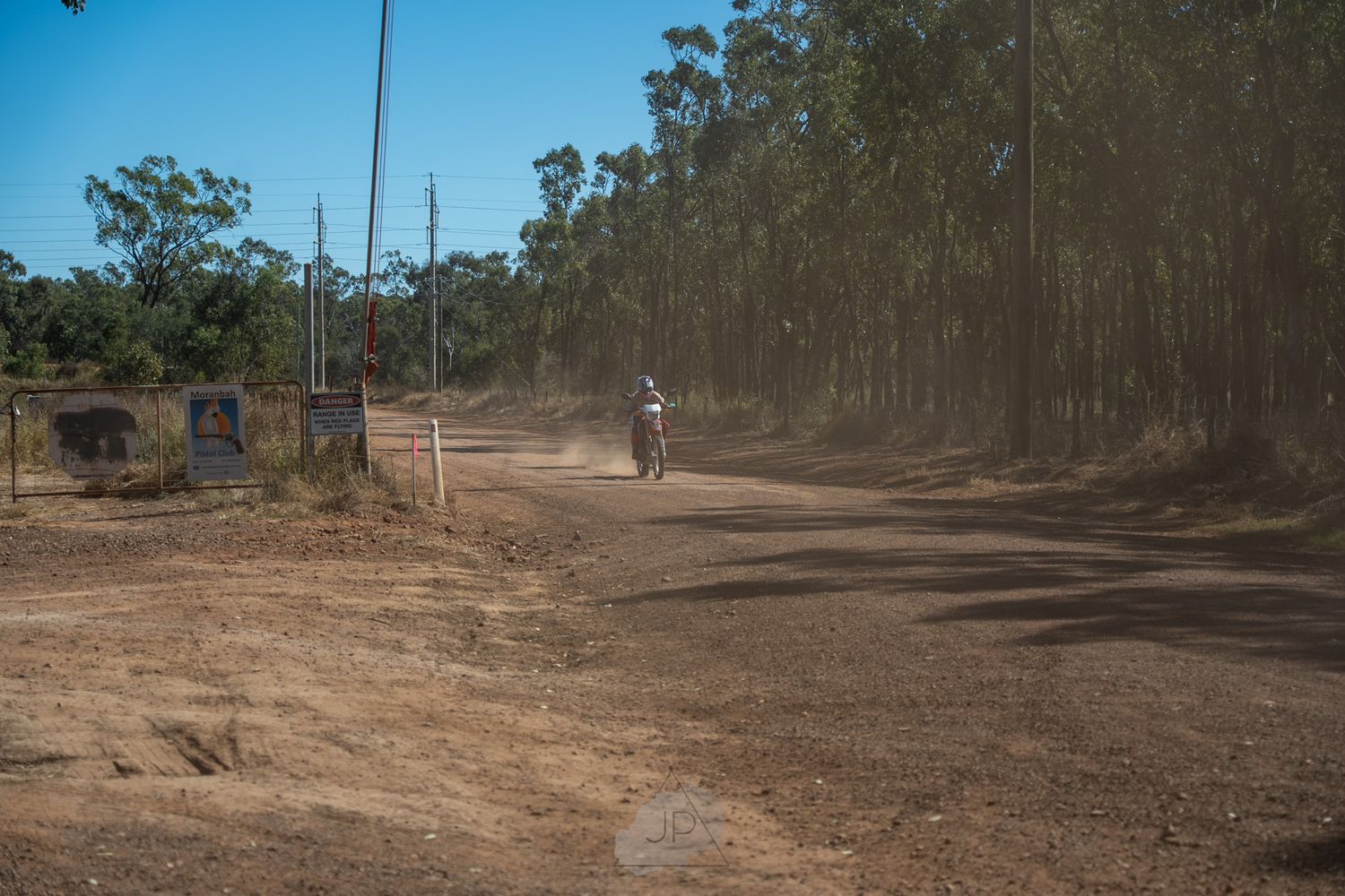 Dirt bike riders navigate through a dusty forest trail on a sunny day.