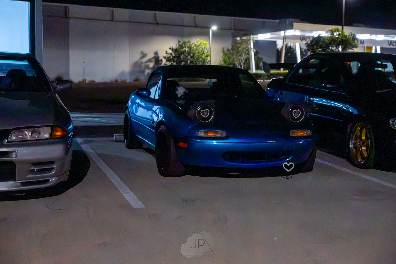 Modified car show at night showing multiple vehicles with custom lighting and modifications in a parking area.