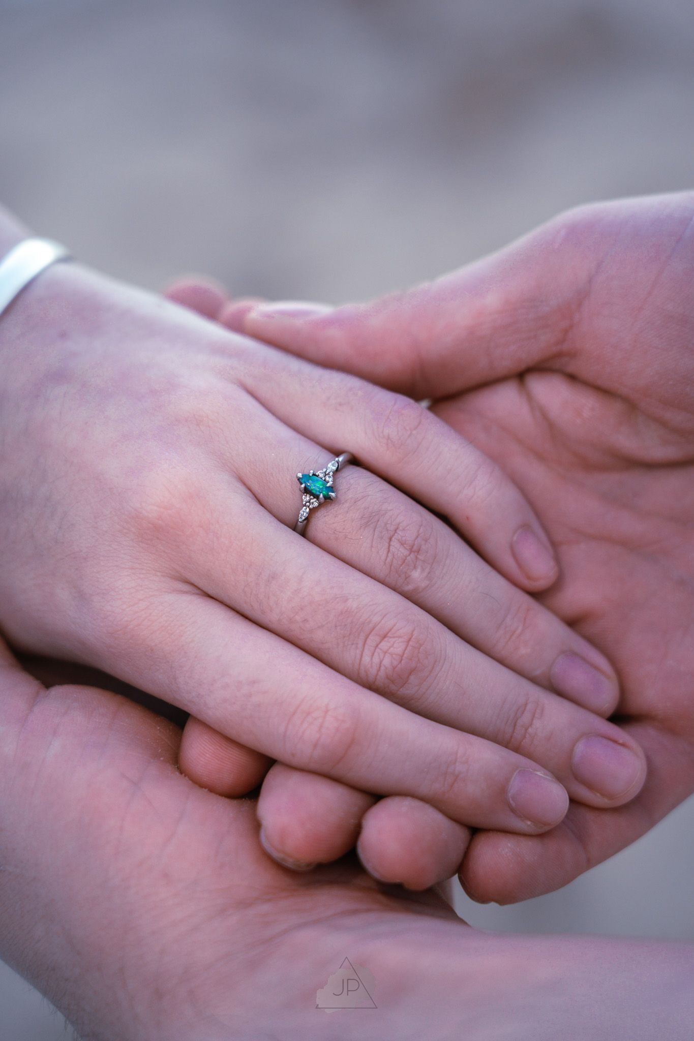 Two hands tenderly intertwined with a delicate blue gemstone engagement ring visible on one finger.