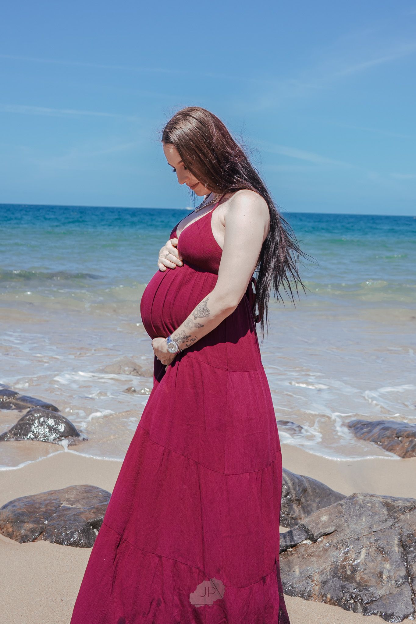 A person in a burgundy dress stands on a rocky beach with turquoise ocean waters and blue skies in the background.