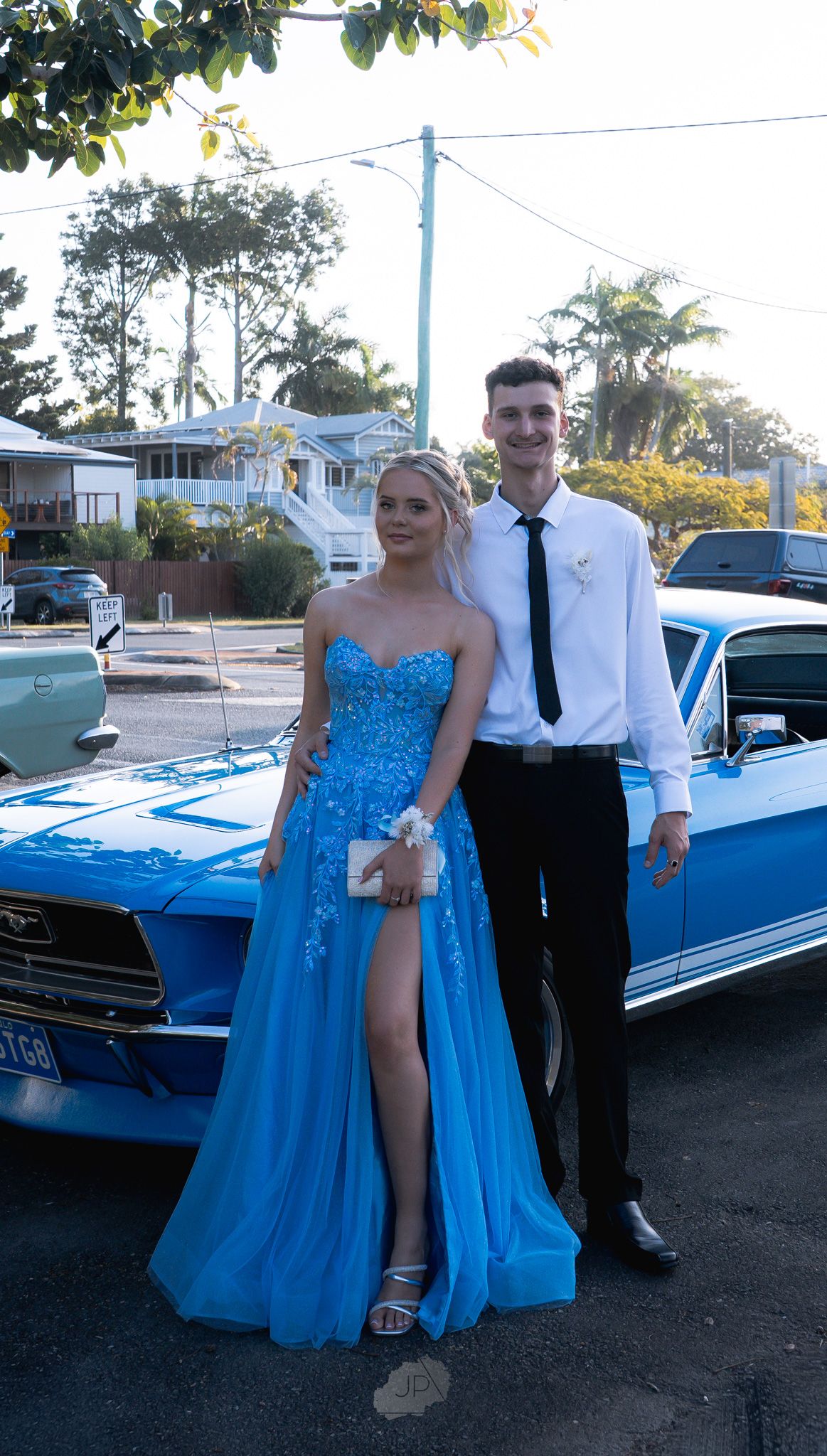 A couple poses in formal attire next to a classic blue car at dusk during what appears to be a prom or special event.