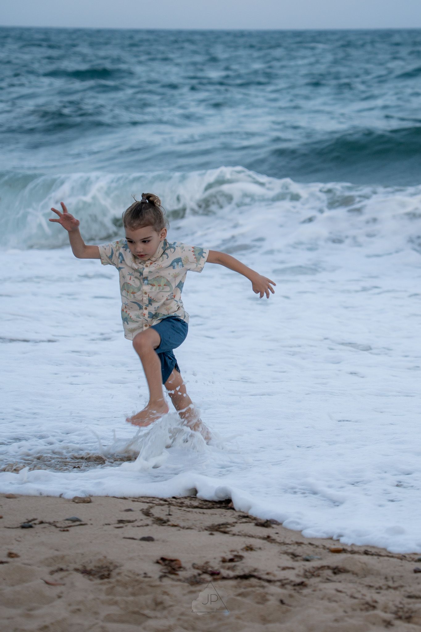 A child in a floral shirt and blue shorts leaps away from incoming ocean waves on a sandy beach.