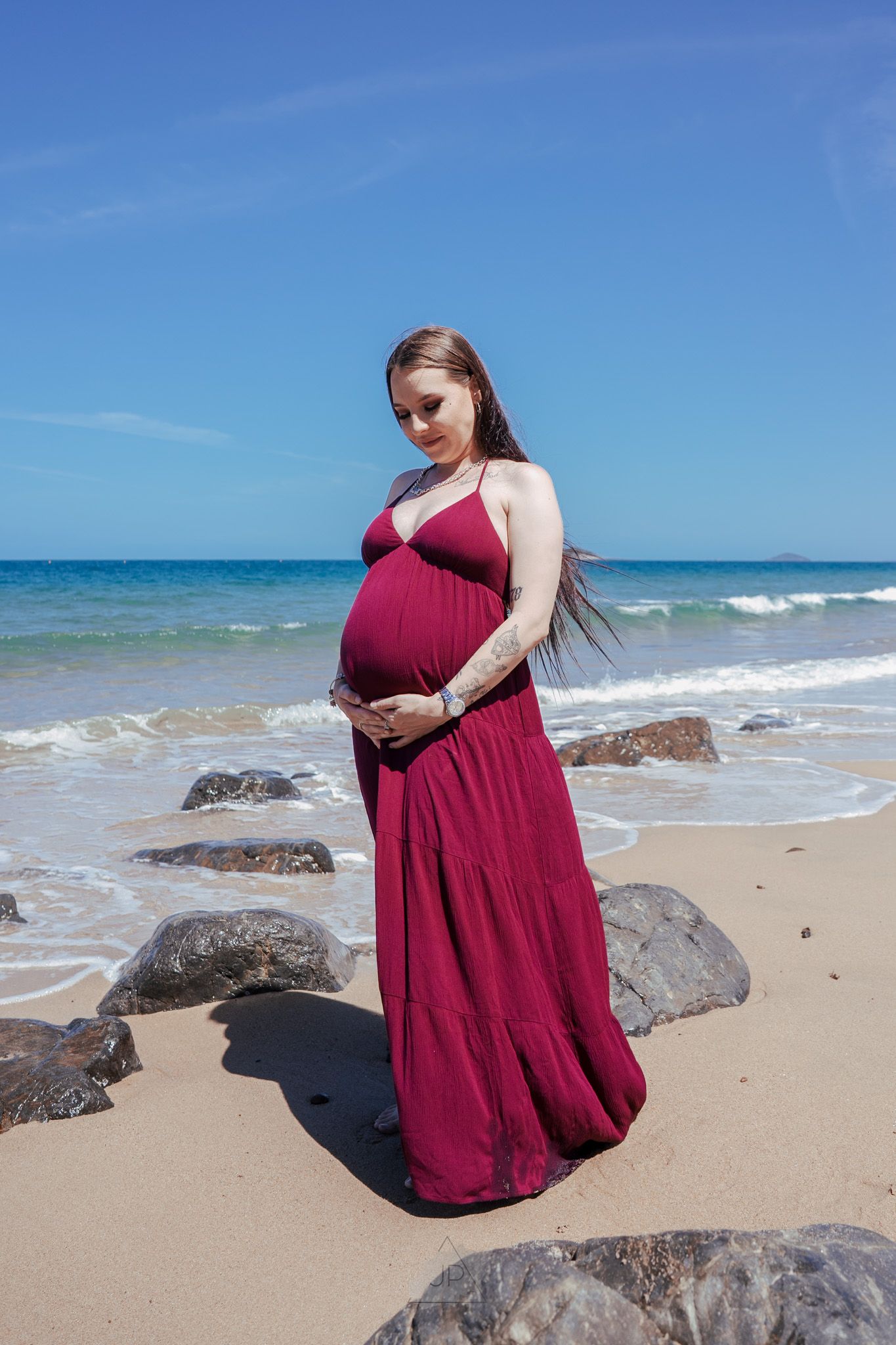 A person in a burgundy dress sits on rocky beach with ocean waves and blue sky in the background.