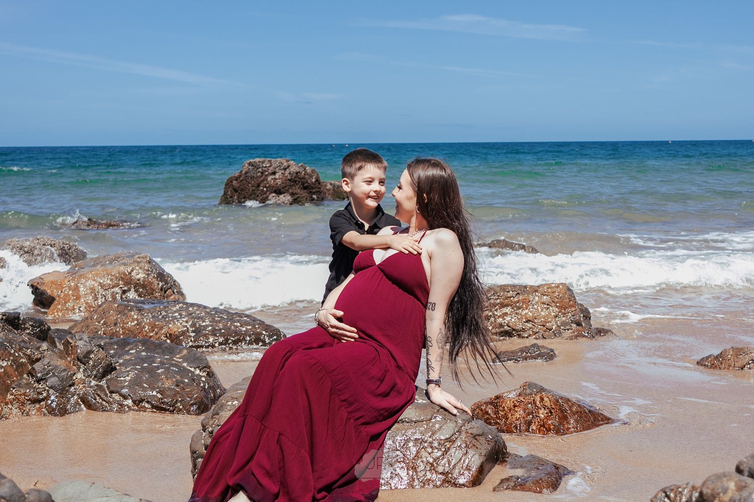 Romantic couple in burgundy outfits embracing on a scenic beach with rocky outcrops and blue ocean waves.