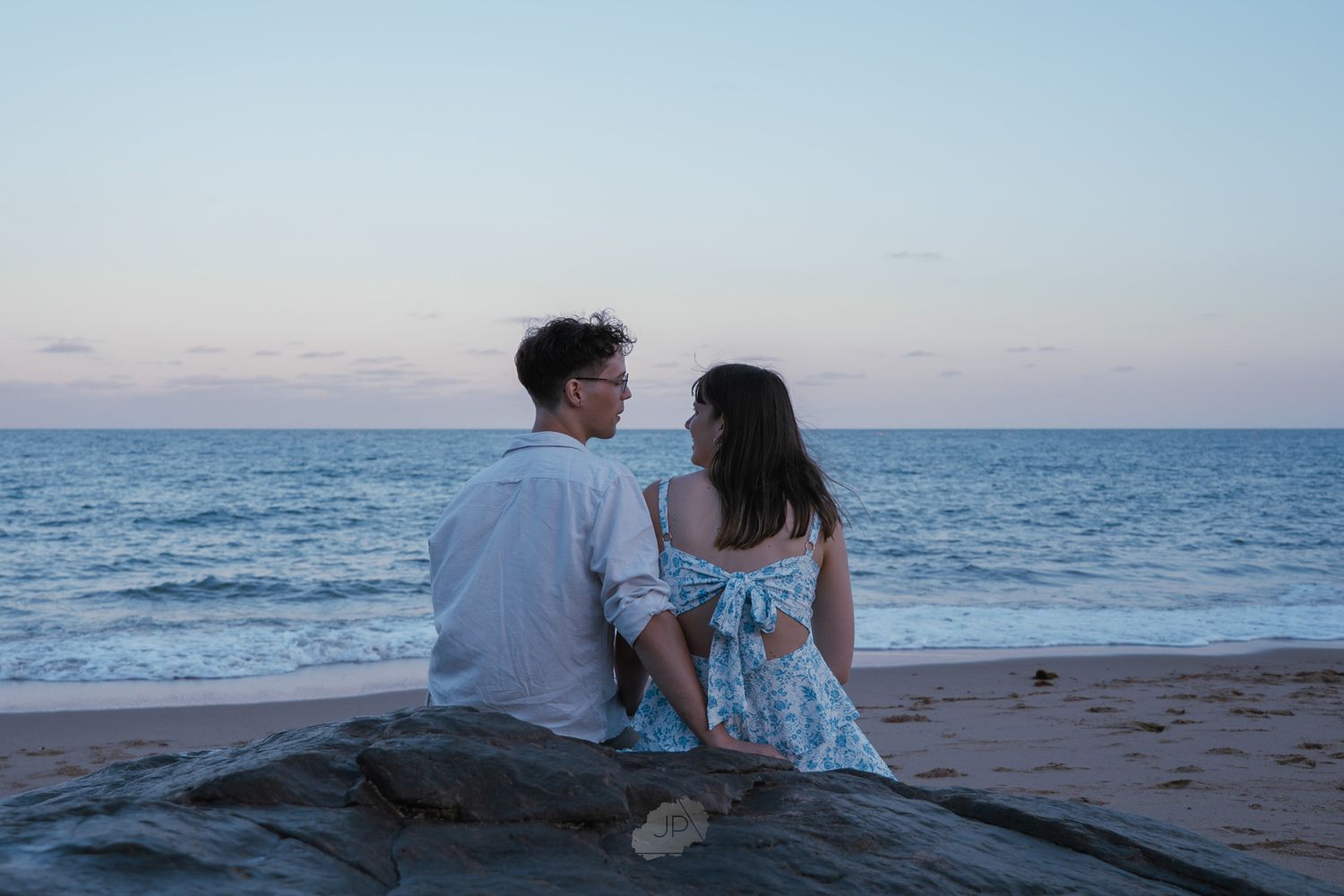 Two people sit together on rocks overlooking a serene ocean view at sunset.