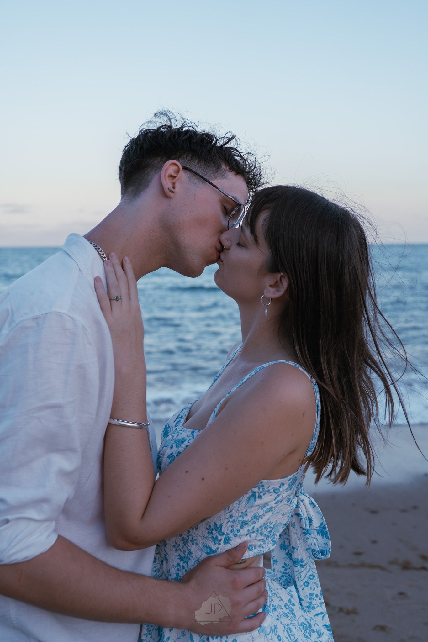 A romantic couple shares intimate kisses on a beach at sunset with ocean waves in the background.