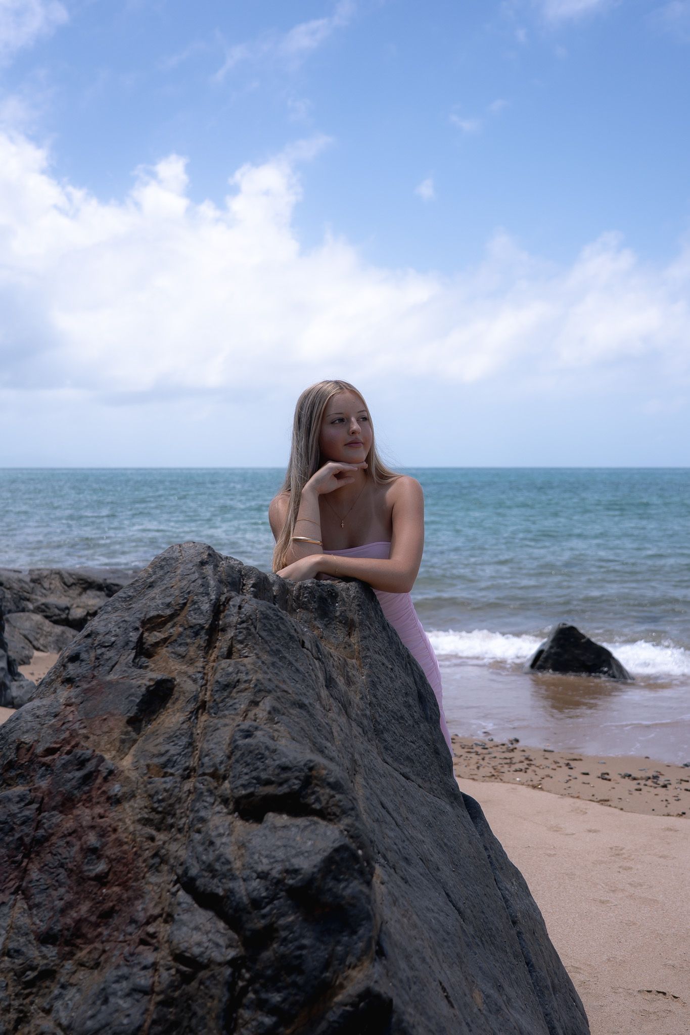 A person lounges on dark volcanic rocks at a scenic beach with turquoise waters and white clouds in the background.