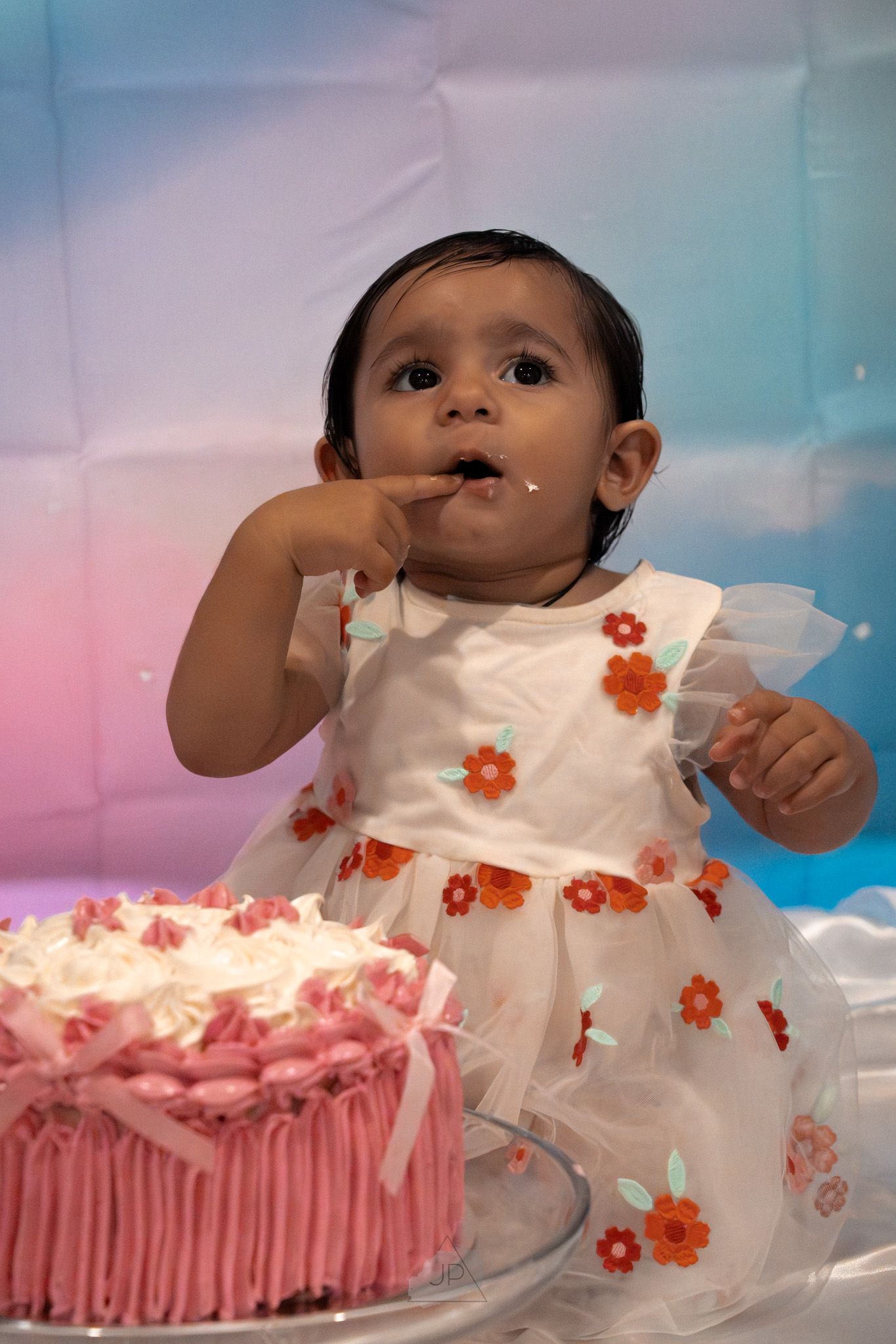 A baby in a white floral dress sits next to a pink birthday cake in a series of progressive photos.