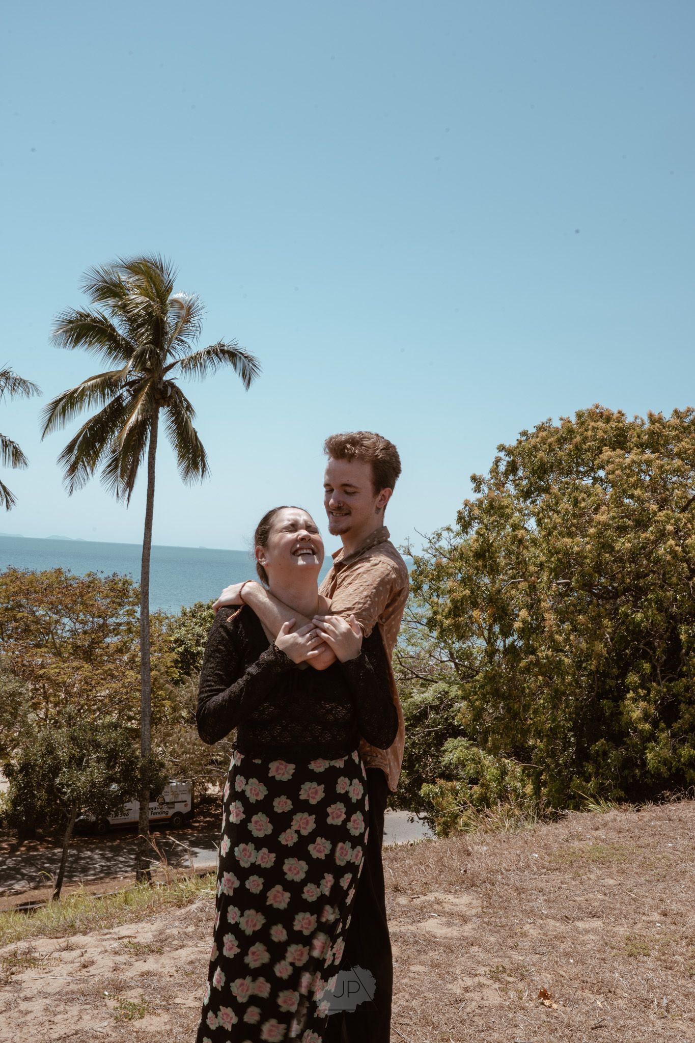 A couple embraces on a sunny hilltop with palm trees and ocean views in the background.