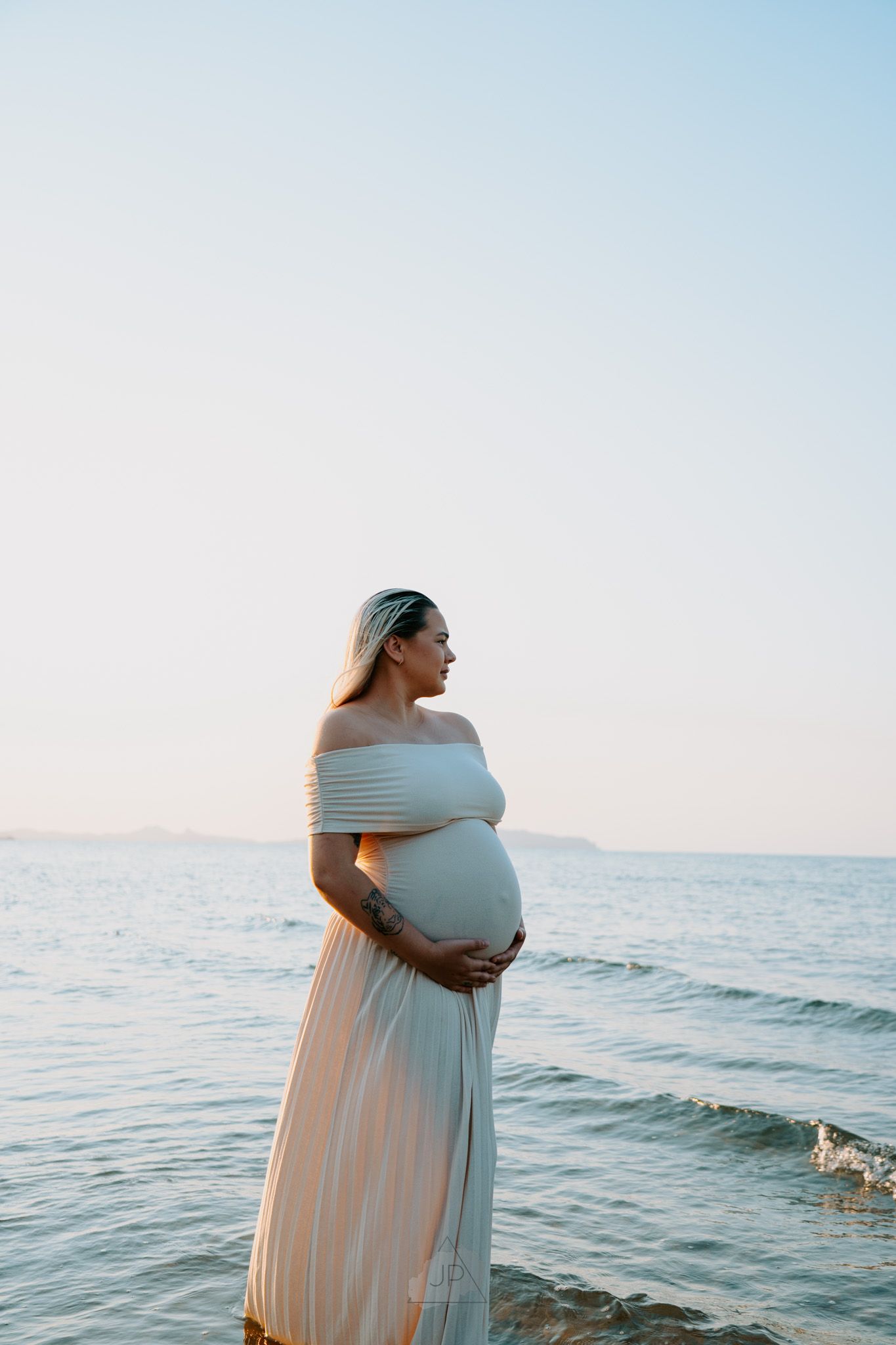 Expectant mother in a flowing white dress stands by the ocean during sunset for a serene maternity photoshoot.