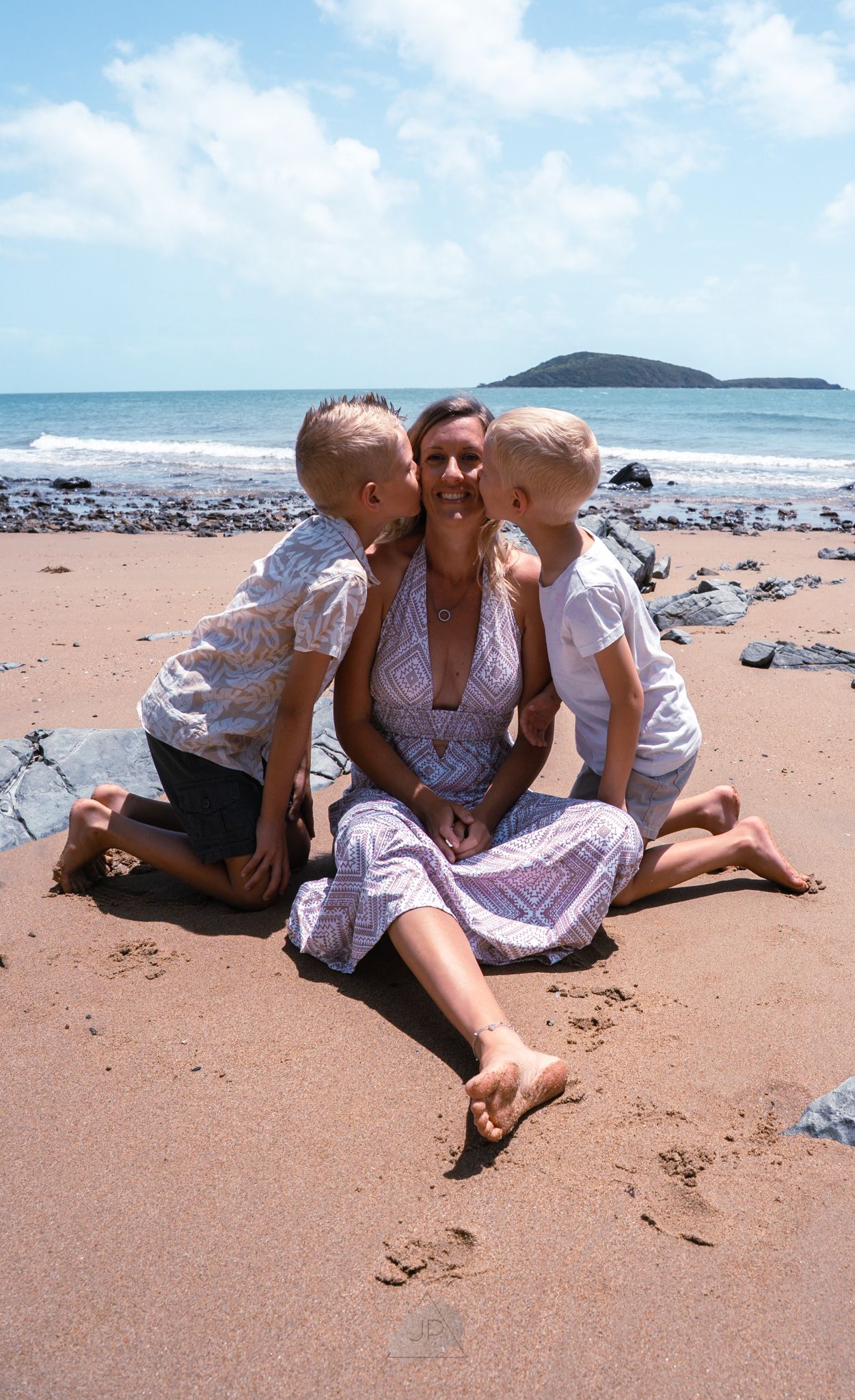Three friends in white dresses relax together on a sunny beach with waves and rocks visible in the background.