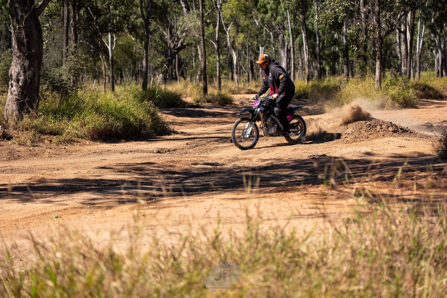 A motorcycle rider navigates a dirt trail through the Australian bush on a sunny day.