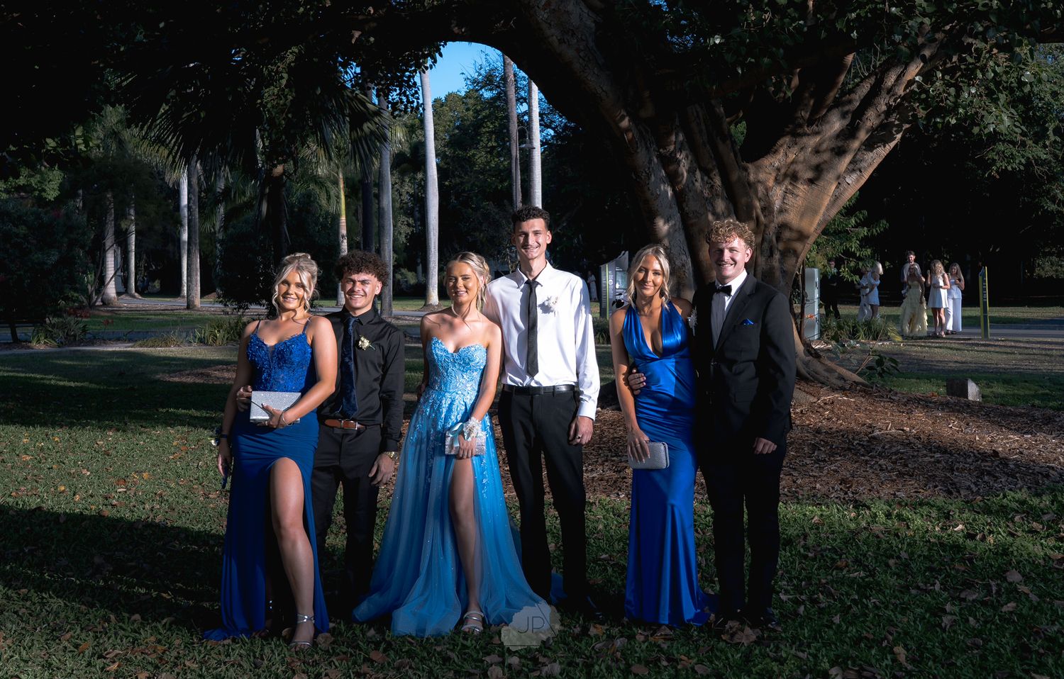 A formal group in blue dresses and suits poses together outdoors under trees in a park setting.