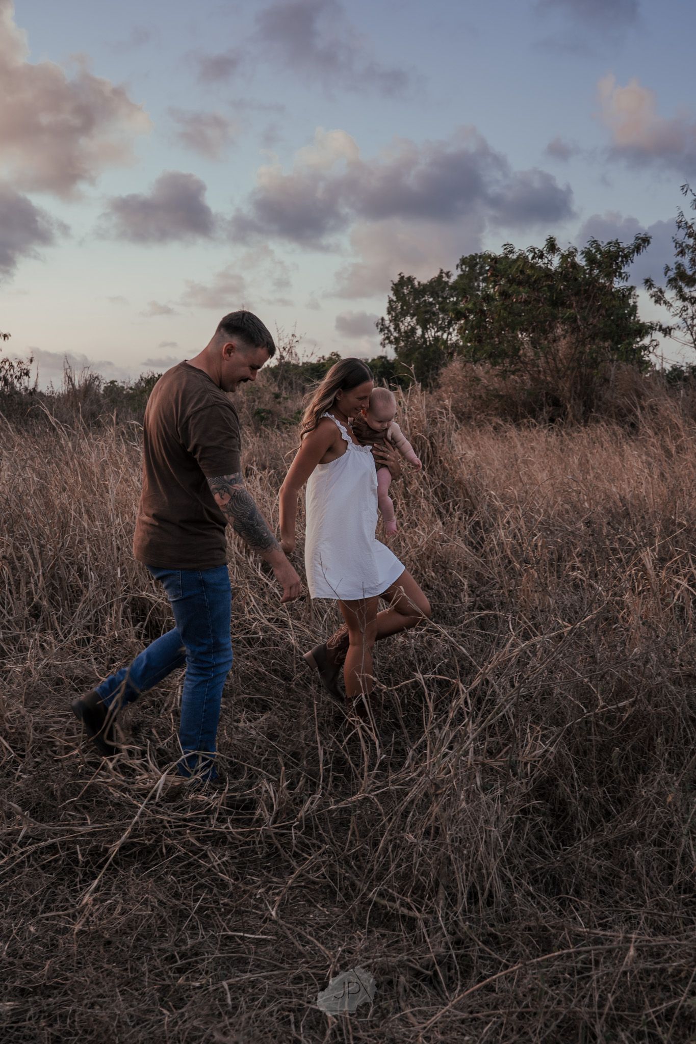 A couple walks together through tall brown grass during sunset in a rustic outdoor setting.