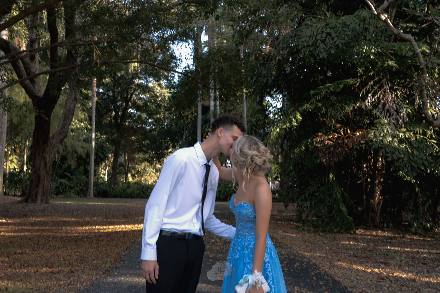 A couple in formal attire share a romantic moment in a tree-lined park during golden hour.