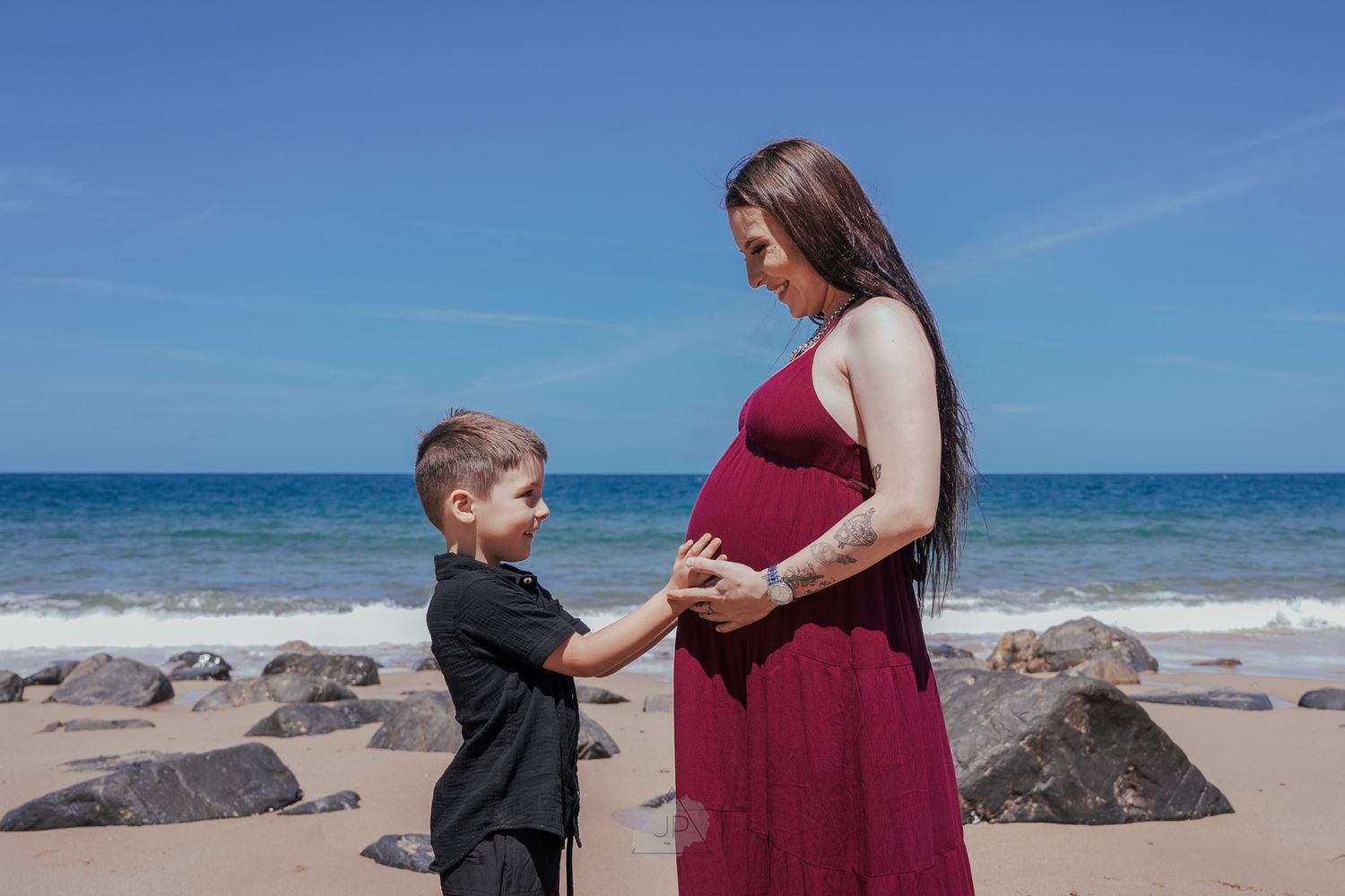 A maternity photo shoot at a scenic beach with large rocks and blue ocean waves in the background.