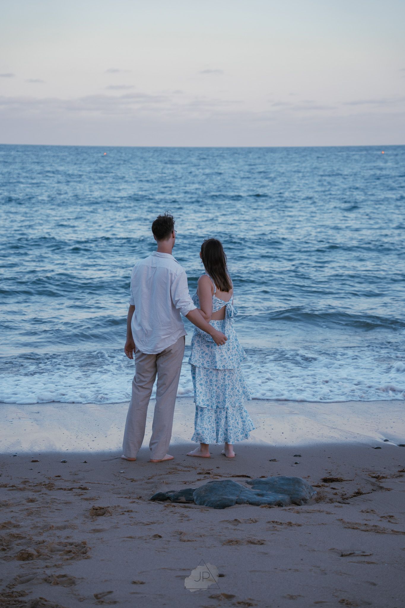A couple stands together on a beach at sunset looking out at the blue ocean waves.