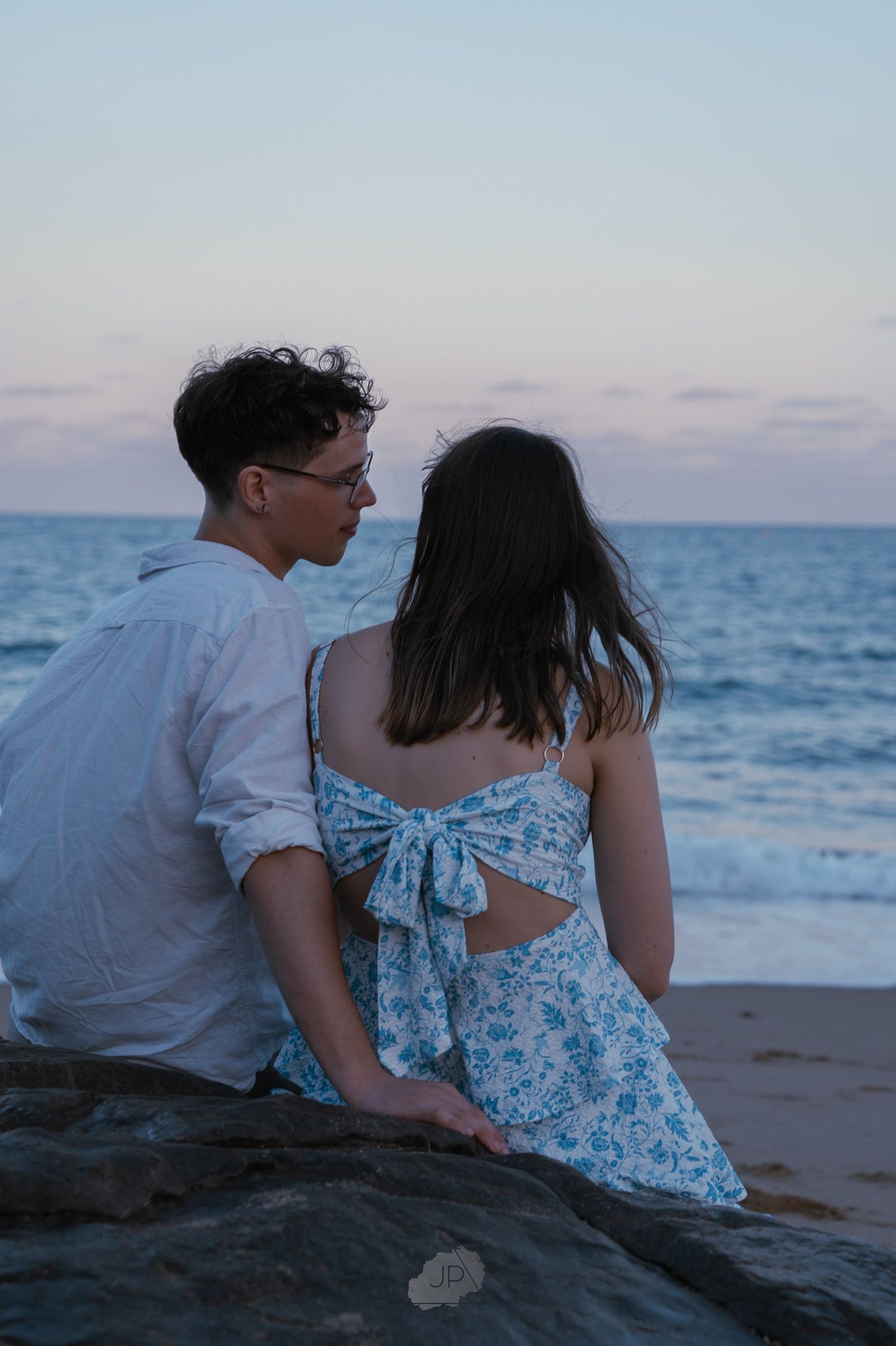 A couple sits together on a rocky beach at sunset overlooking the ocean in a romantic moment.