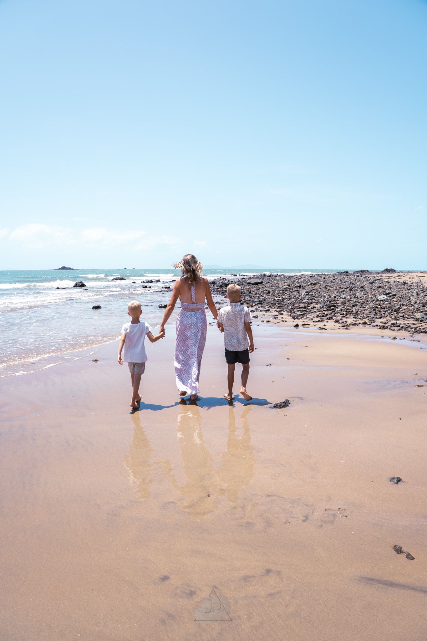 Three people walking together along a scenic sandy beach shoreline with gentle waves and rocky outcrops in the distance.