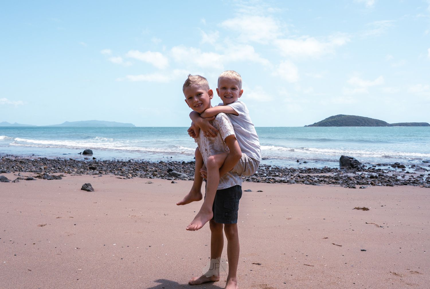 Family members play and walk together on a scenic beach with rocky coastline and blue water in the background.