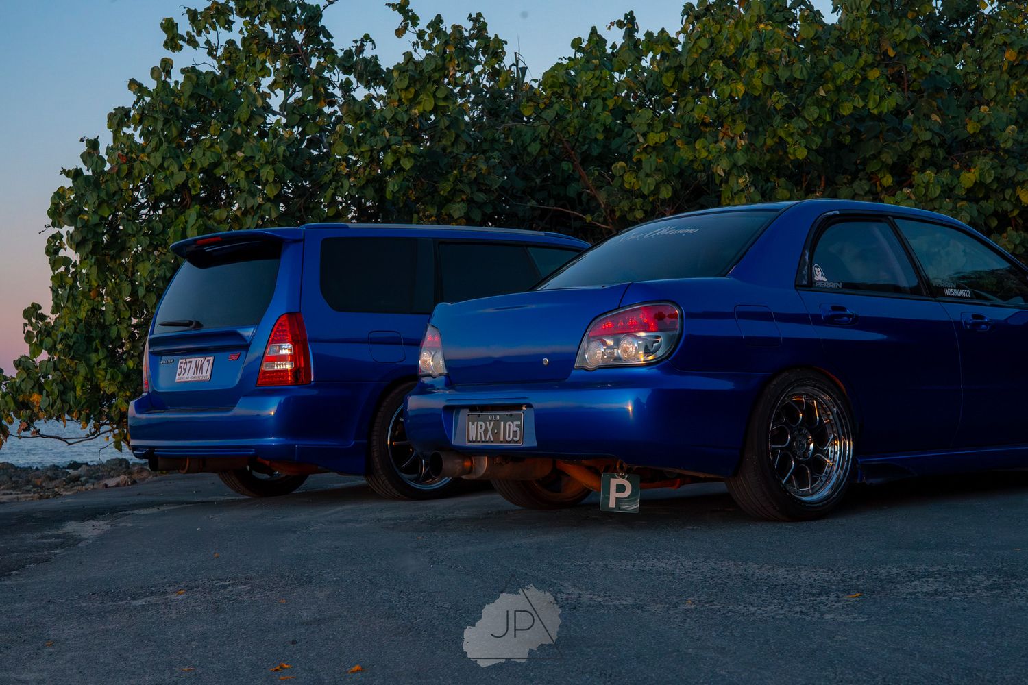 A blue Volvo wagon and blue Subaru WRX parked together at dusk with trees in the background.