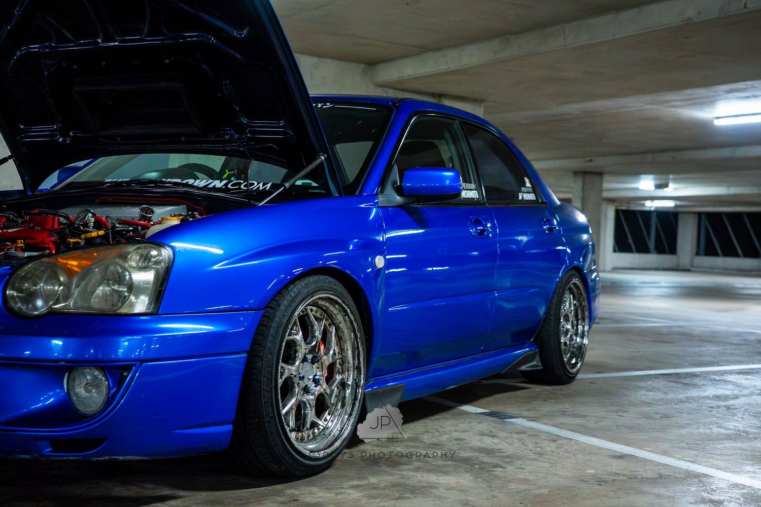 Custom lowered Subaru WRX in metallic blue showcasing aftermarket wheels in underground parking structure.