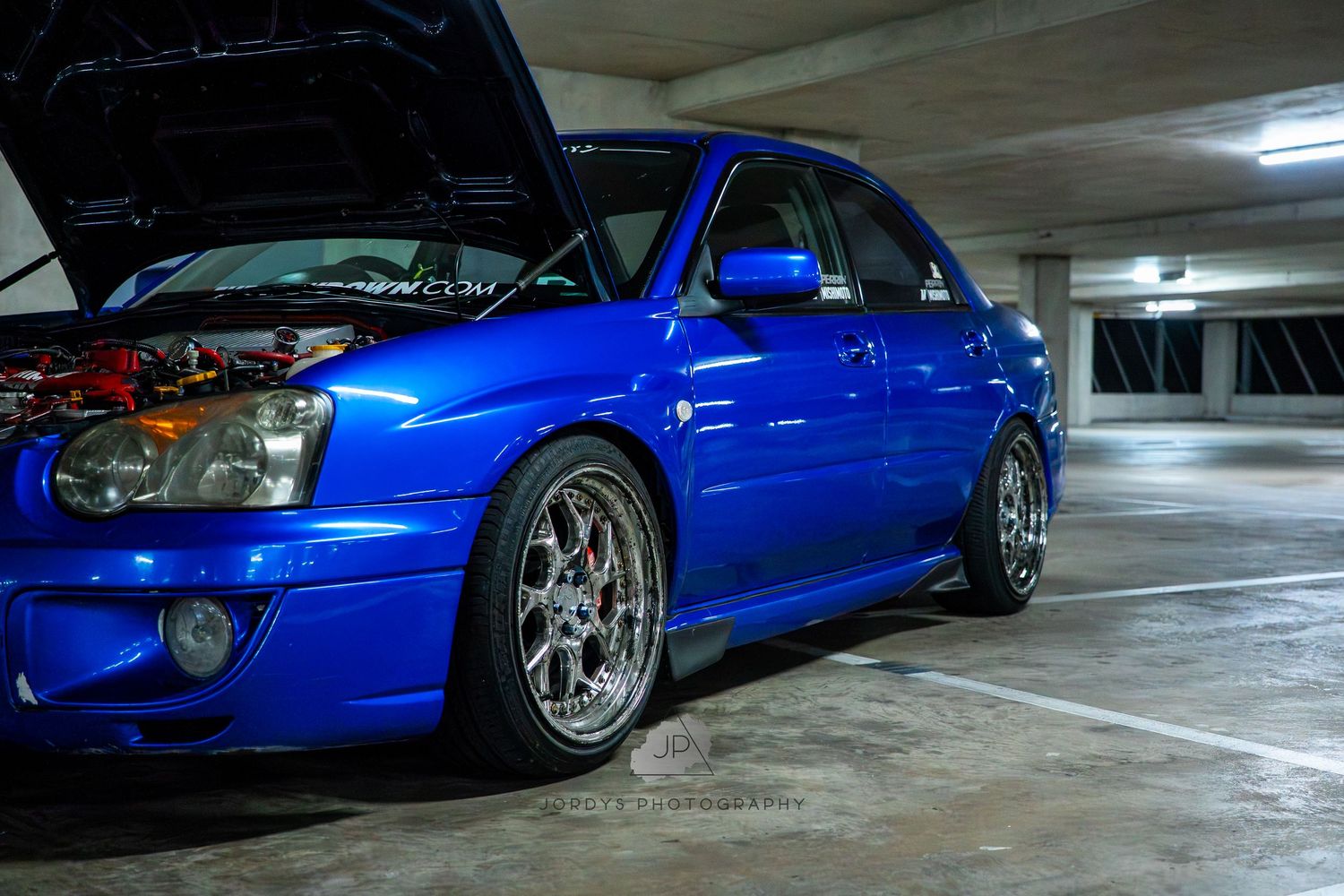 A bright blue modified sports car with custom wheels and low stance parked in a dimly lit garage or parking area.