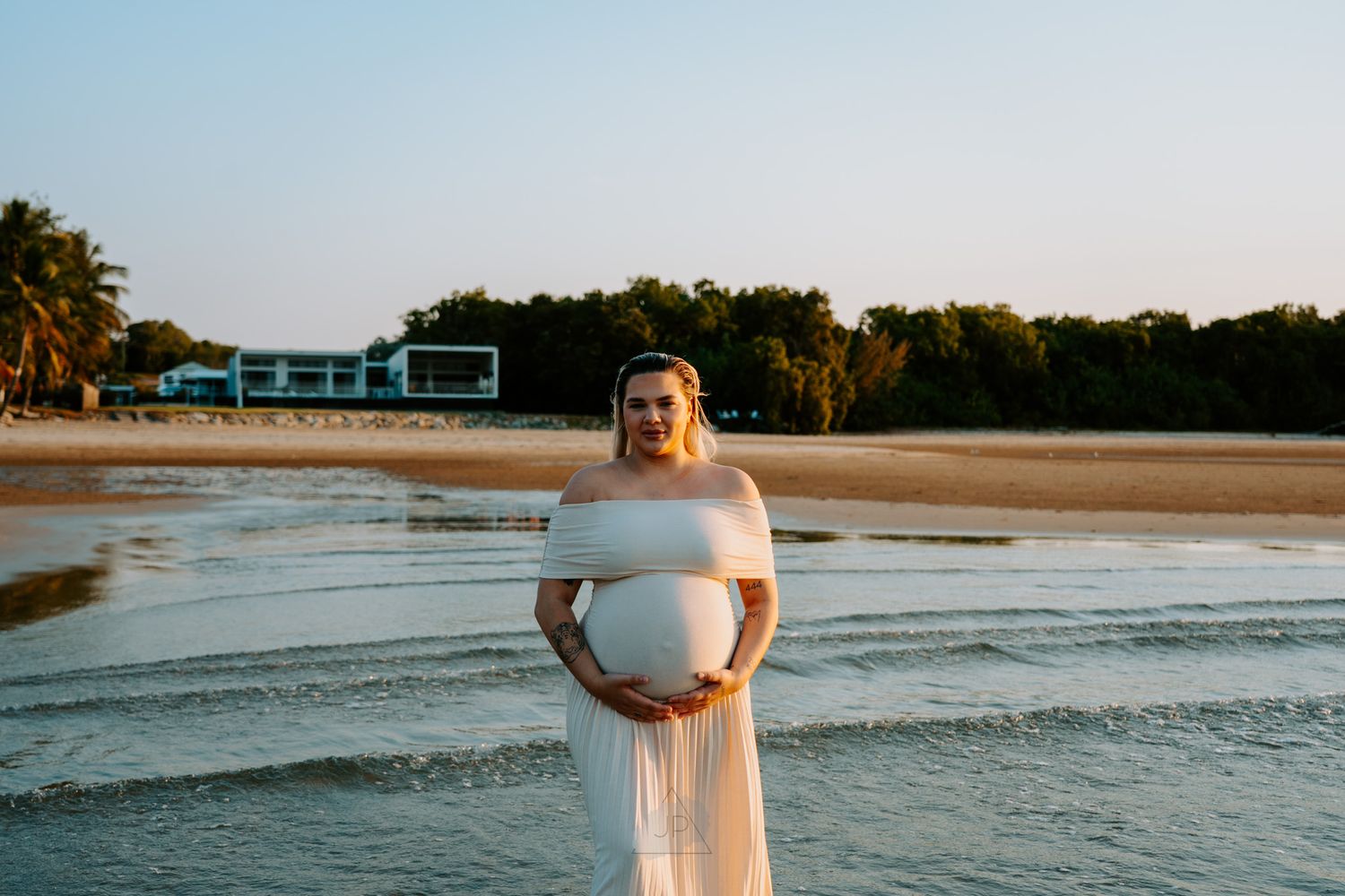 A person in a white dress walks along a serene beach at sunset with gentle waves rolling onto the shore.